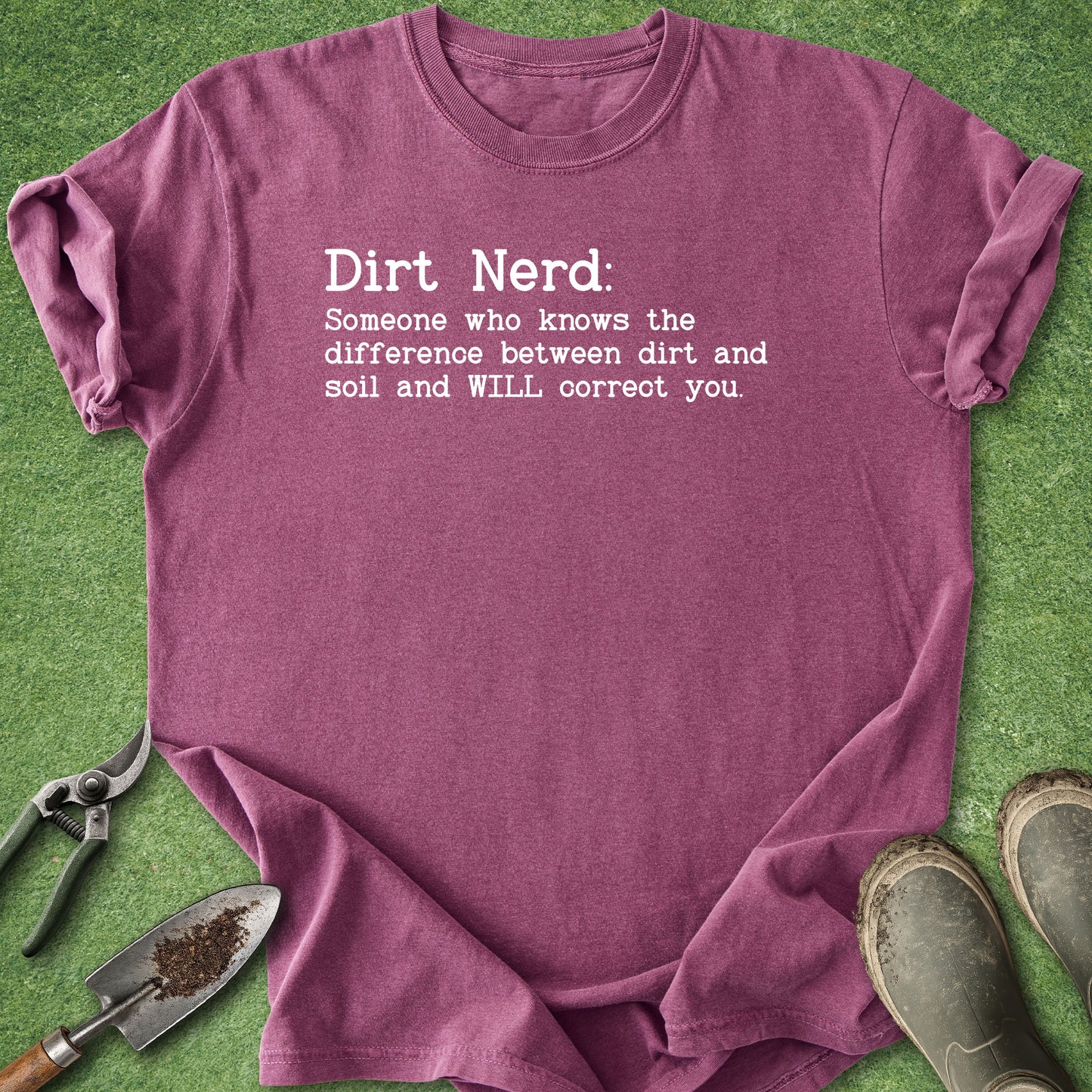 A heathered mauve short-sleeve t-shirt with white printed text reading 'Dirt Nerd: Someone who knows the difference between dirt and soil and WILL correct you.' laid flat on green grass-like material, with gardening tools and boots around it.
