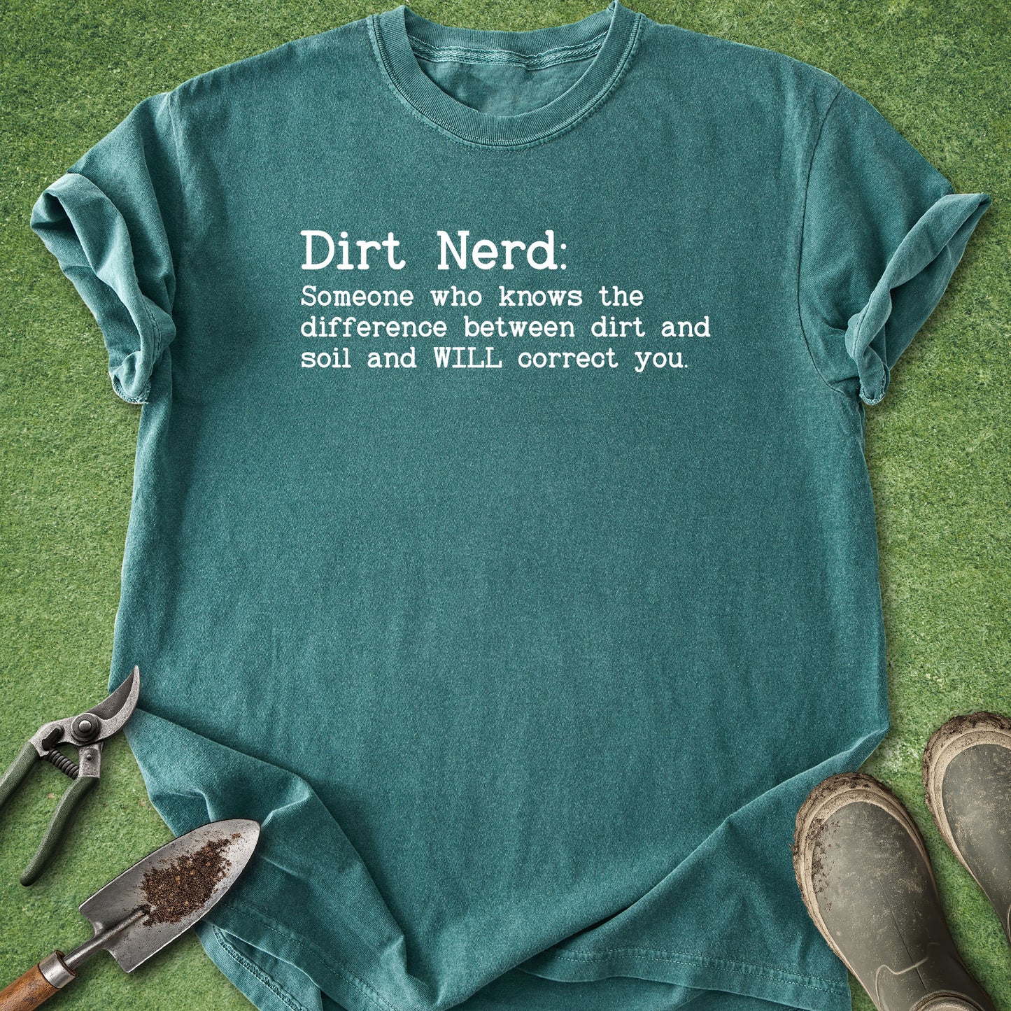 A teal graphic t-shirt laid flat on green grass with gardening props, including pruning shears, a trowel, and muddy boots. The t-shirt features white text that reads 'Dirt Nerd: Someone who knows the difference between dirt and soil and WILL correct you.