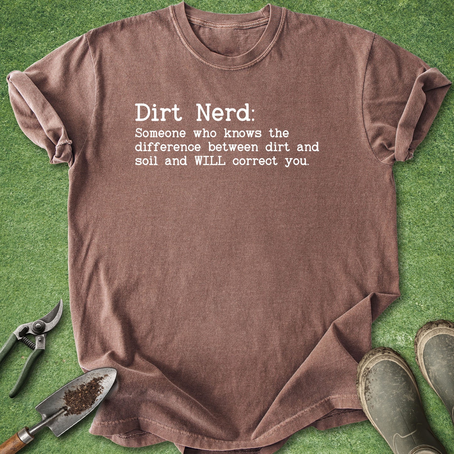 A brown short-sleeve graphic T-shirt laid flat on green grass-like material, with gardening tools and muddy boots around it. The shirt reads, 'Dirt Nerd: Someone who knows the difference between dirt and soil and WILL correct you.