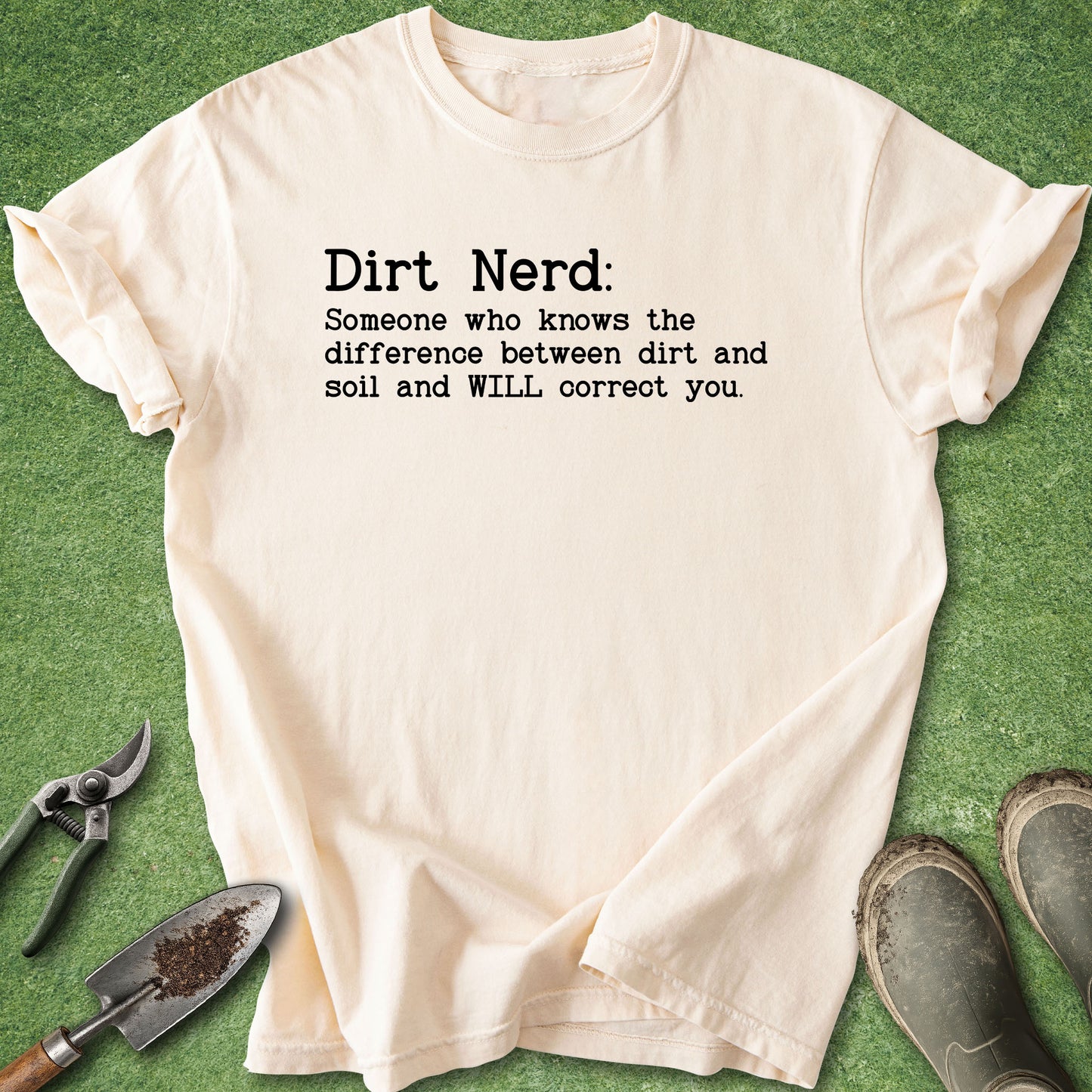 A cream-colored t-shirt with black printed text reading 'Dirt Nerd: Someone who knows the difference between dirt and soil and WILL correct you.' is laid flat on green grass with gardening tools and muddy boots around it.