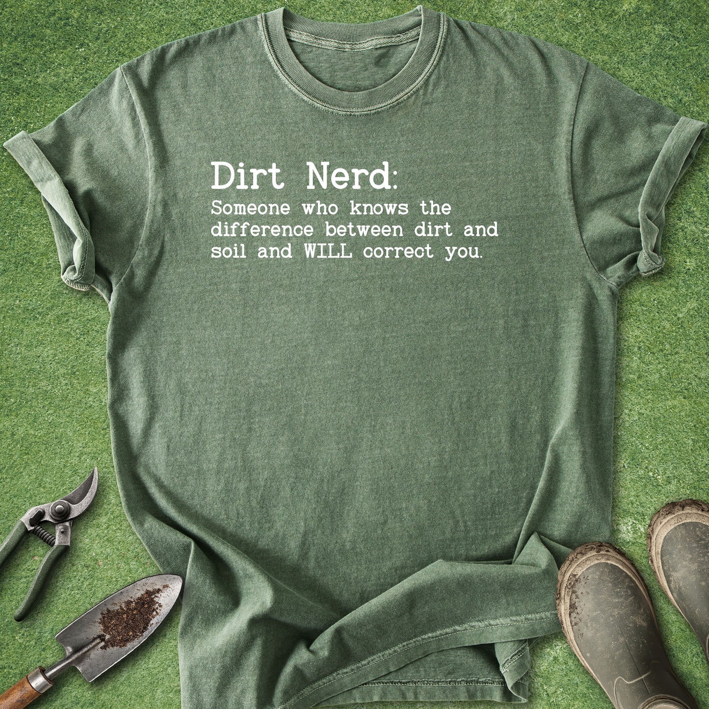 An olive green graphic t-shirt laid flat on a green grass-like surface, with gardening tools and muddy boots around it. The shirt has white printed text that reads: 'Dirt Nerd: Someone who knows the difference between dirt and soil and WILL correct you.
