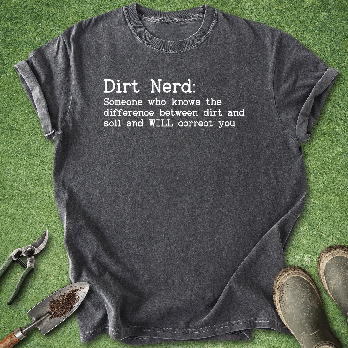 A dark charcoal gray t-shirt with a white printed graphic reading 'Dirt Nerd: Someone who knows the difference between dirt and soil and WILL correct you.' The shirt is laid flat on a green grass-like surface, surrounded by gardening props including pruning shears, a trowel, and muddy boots.