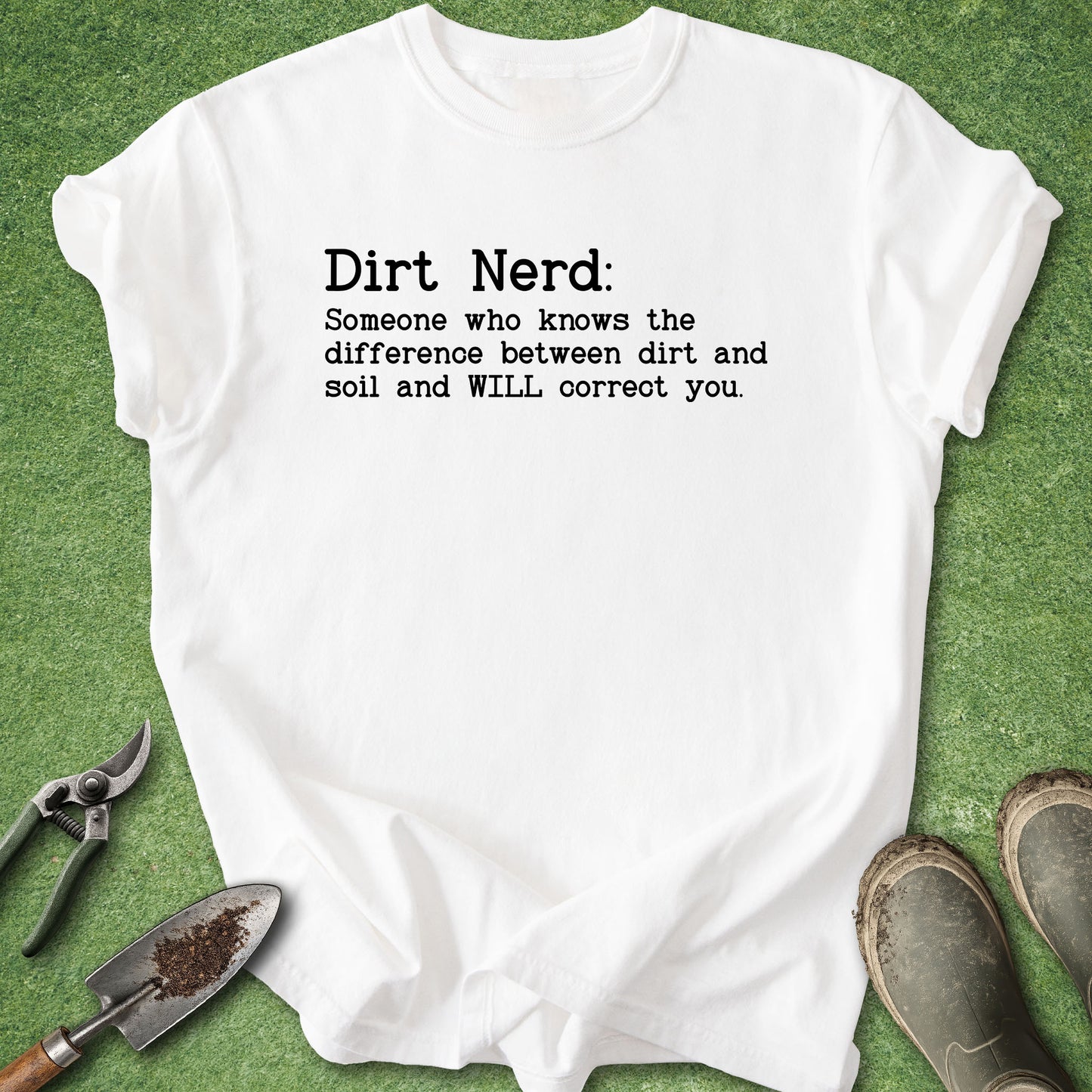 A white graphic t-shirt with black text reading 'Dirt Nerd: Someone who knows the difference between dirt and soil and WILL correct you.' is laid flat on green artificial grass with gardening props including pruning shears, a trowel, and boots.