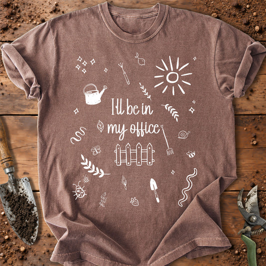 A brown short-sleeve t-shirt with a white graphic print that reads 'I'll be in my office' surrounded by gardening and office-themed doodles, laid flat on a rustic wooden surface with gardening tools.