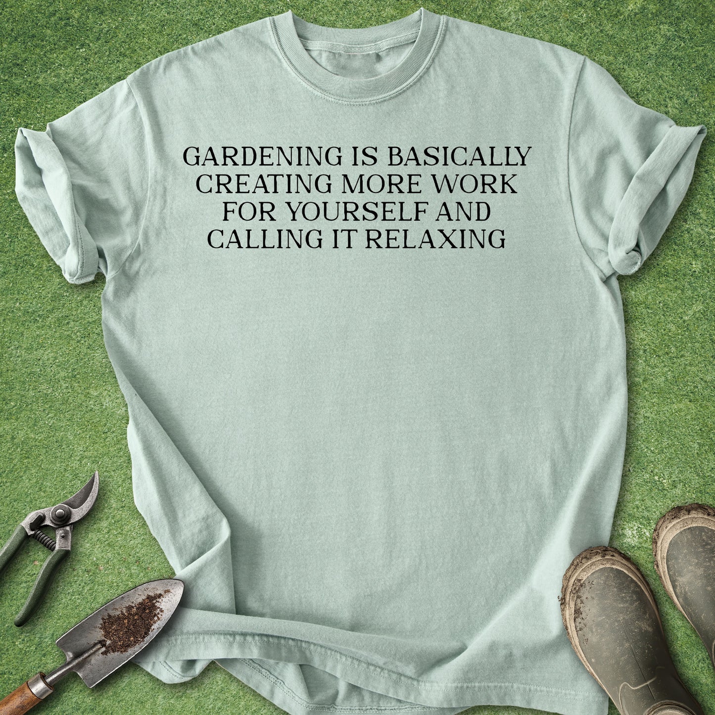 A light sage green t-shirt with black printed text reading 'GARDENING IS BASICALLY CREATING MORE WORK FOR YOURSELF AND CALLING IT RELAXING', laid flat on green grass with gardening tools (trowel, pruning shears) and boots nearby.
