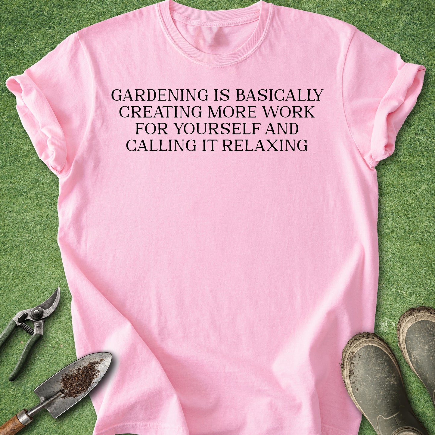 A light pink short-sleeve t-shirt with black printed text reading 'GARDENING IS BASICALLY CREATING MORE WORK FOR YOURSELF AND CALLING IT RELAXING', laid flat on green grass with gardening tools and boots around it.