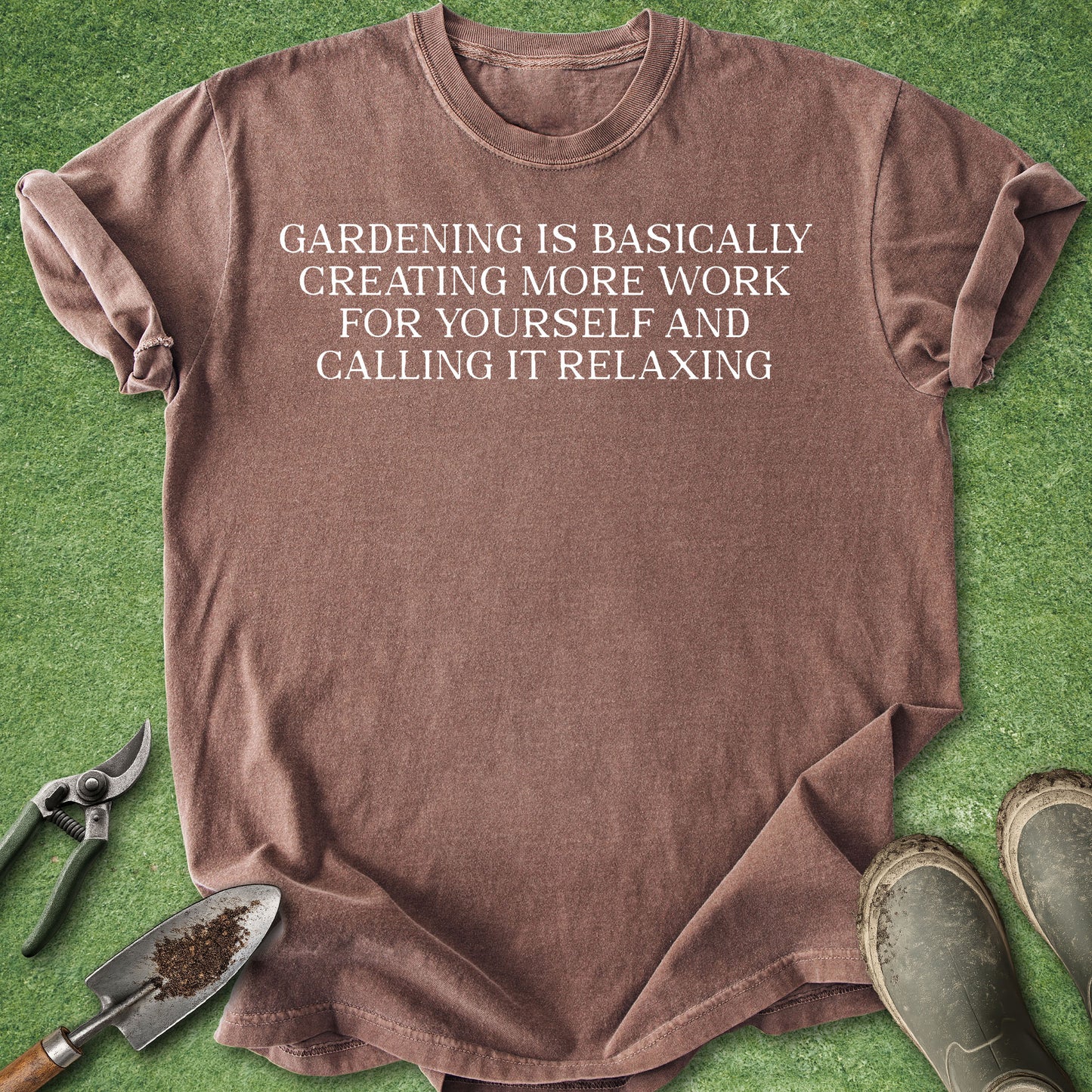 A brown short-sleeve t-shirt with white text reading 'GARDENING IS BASICALLY CREATING MORE WORK FOR YOURSELF AND CALLING IT RELAXING' is laid flat on green grass-like material with gardening tools and boots around it.