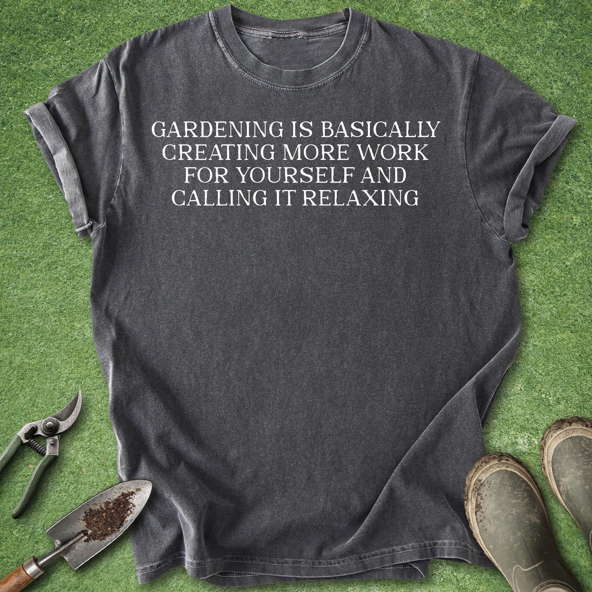 A dark charcoal gray t-shirt with the white printed text 'my retirement plans' and simple gardening icons (plant, shears, watering can) on the chest. The shirt is laid flat on green grass with gardening props (trowel, soil, pruning shears, boots) around it.