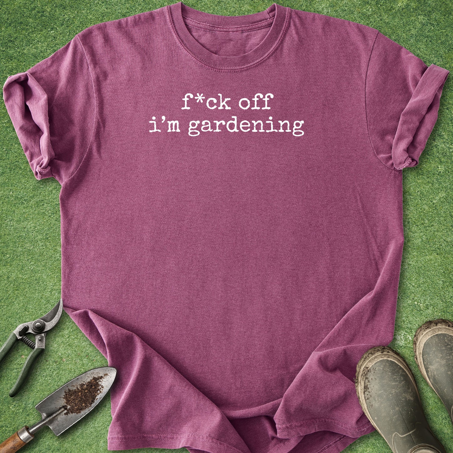 A heathered mauve-purple t-shirt with the white text 'f*ck off i'm gardening' printed on the chest, styled in a flat-lay on green grass with gardening tools (trowel, pruning shears) and boots.
