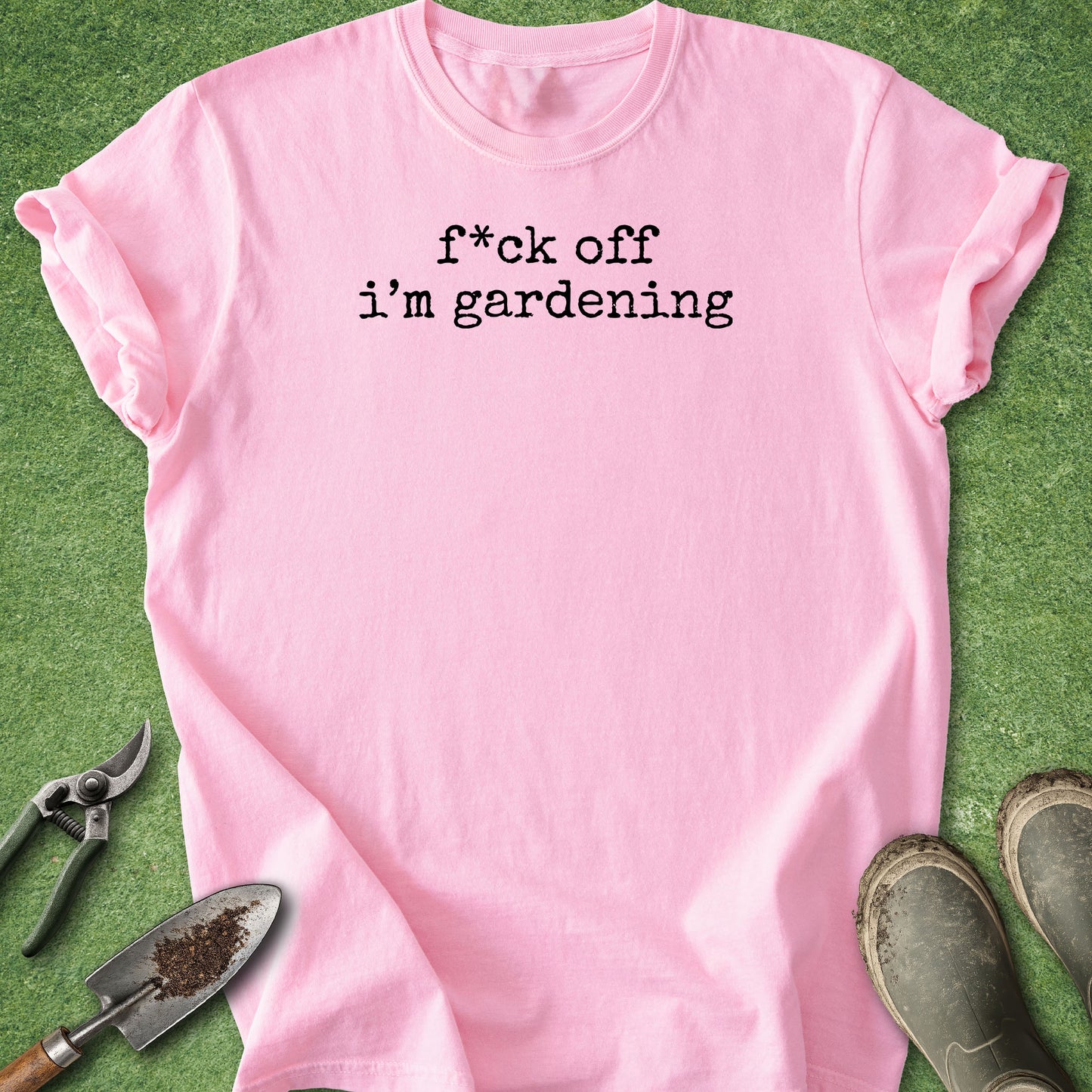 A light pink short-sleeve t-shirt with the black printed text 'f*ck off i'm gardening' laid flat on green grass, styled with gardening tools and boots around it.