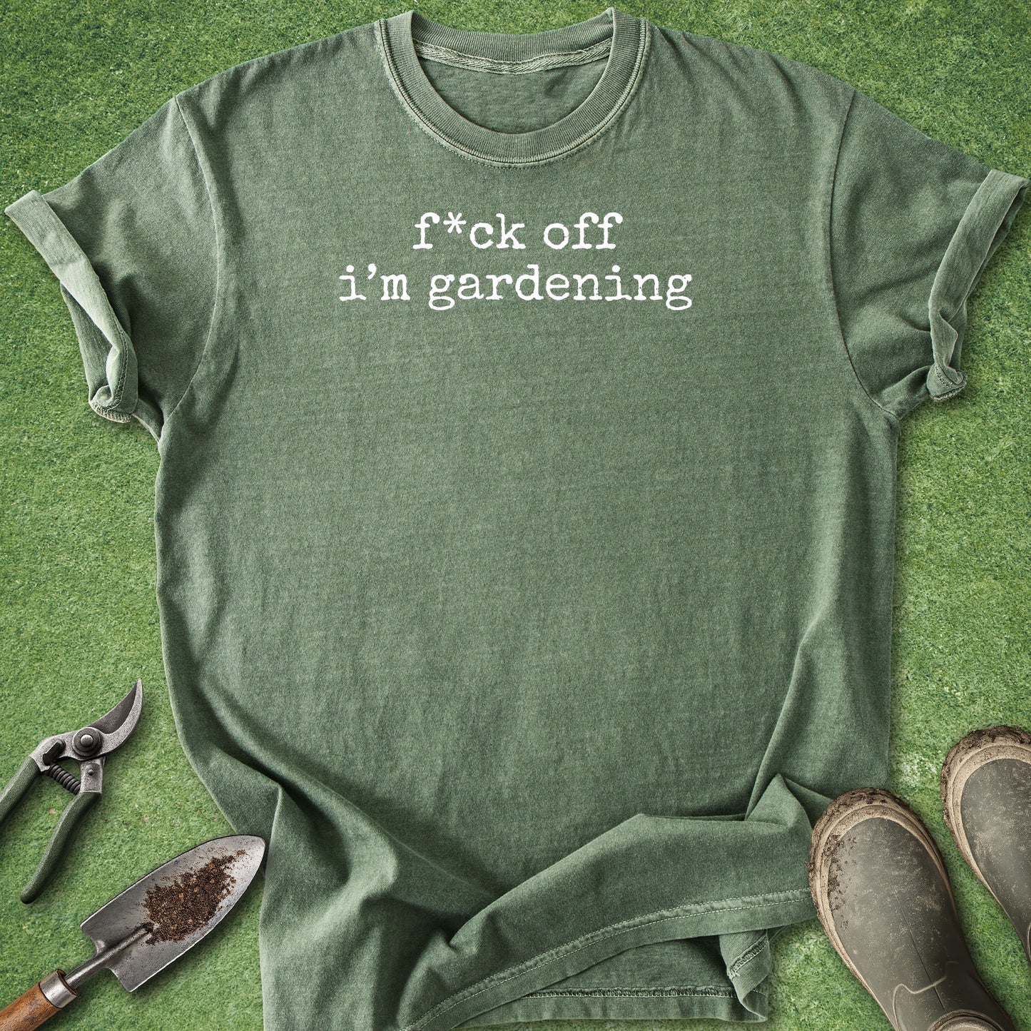 An olive green graphic t-shirt with the text 'f*ck off i'm gardening' printed in white typewriter font, styled in a flat lay on artificial green grass with gardening props including a trowel, pruning shears, and boots.