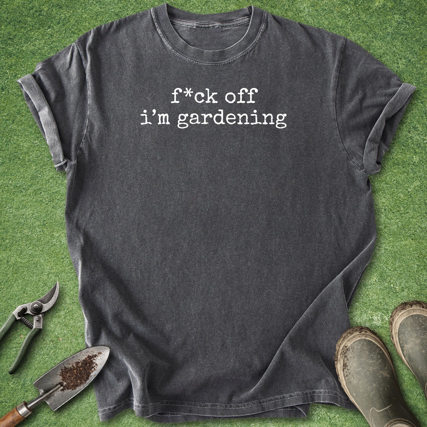A dark charcoal gray t-shirt with the text 'f*ck off i'm gardening' printed in white typewriter font, laid flat on green grass with gardening props including a trowel, pruning shears, and boots.