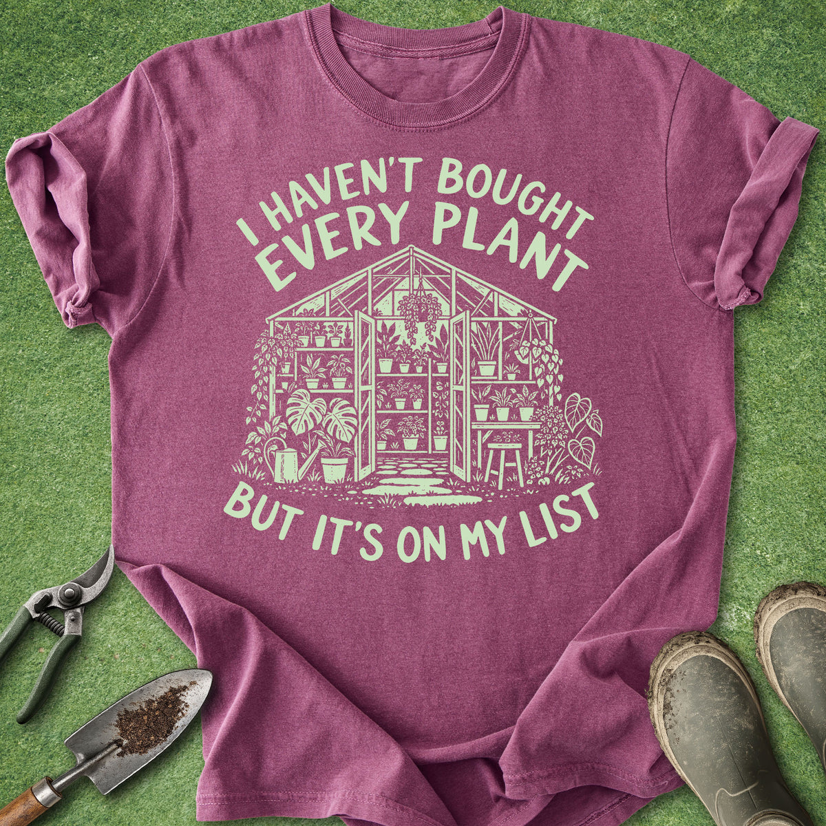 A heathered purple short-sleeve t-shirt with a cream-colored graphic of a greenhouse and the text 'I HAVEN'T BOUGHT EVERY PLANT BUT IT'S ON MY LIST'. The shirt is laid flat on green grass with gardening props including pruning shears, a trowel, and boots.