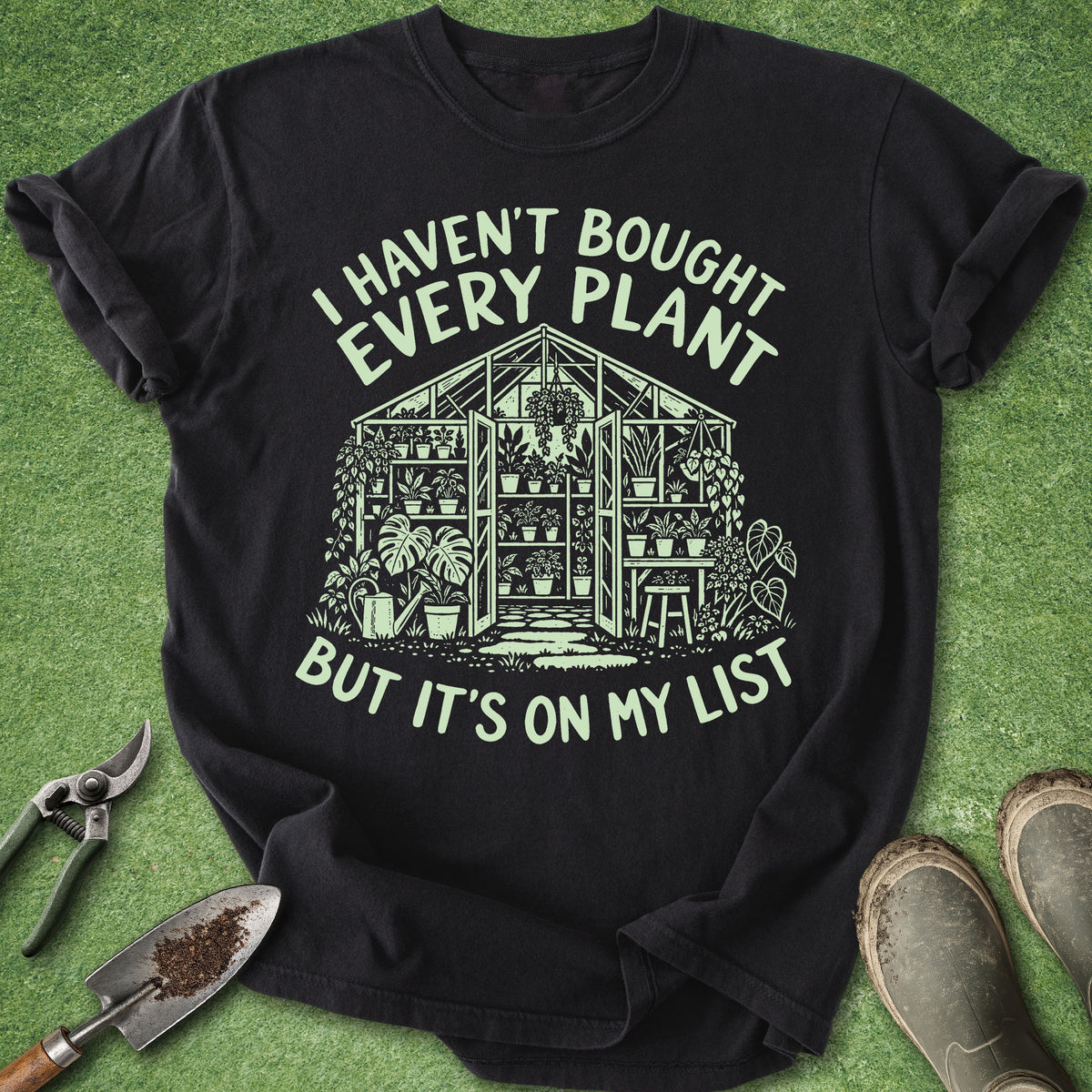 A black graphic t-shirt laid flat on green grass-like artificial turf, with gardening props around it: pruning shears, a trowel, and muddy boots. The shirt has a large light green illustration of a greenhouse and the text 'I HAVEN'T BOUGHT EVERY PLANT BUT IT'S ON MY LIST'.