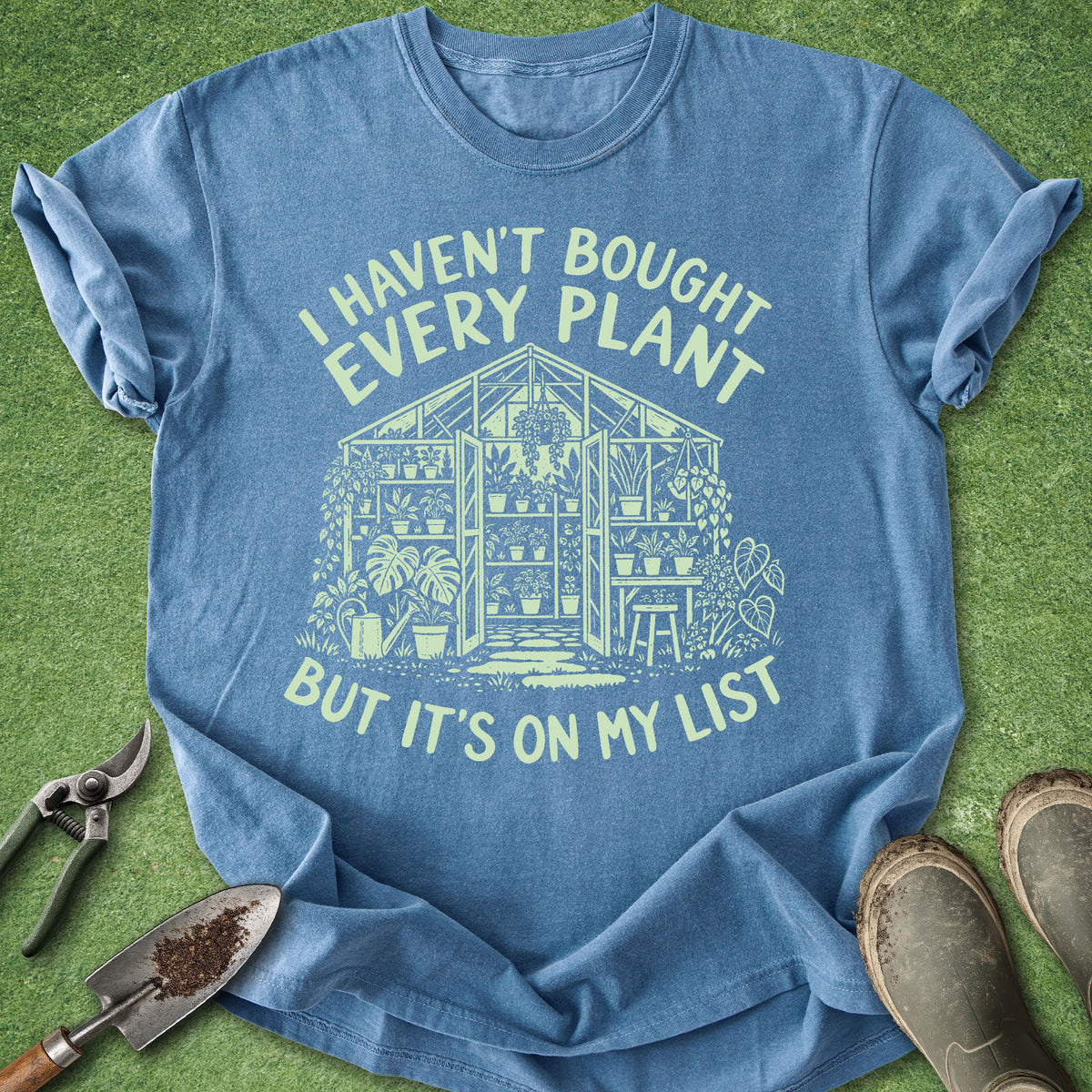A blue graphic t-shirt laid flat on a green grass-like surface, with gardening tools and boots around it. The shirt has a pale yellow-green printed illustration of a greenhouse and the text 'I HAVEN'T BOUGHT EVERY PLANT BUT IT'S ON MY LIST'.