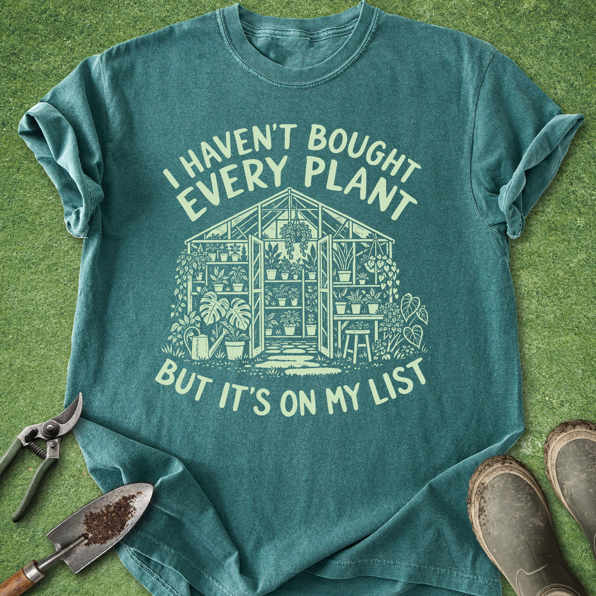 A teal short-sleeve graphic T-shirt laid flat on a green grass-like surface, with gardening tools and boots around it. The shirt has a large cream print of a greenhouse and the text, 'I HAVEN'T BOUGHT EVERY PLANT BUT IT'S ON MY LIST.