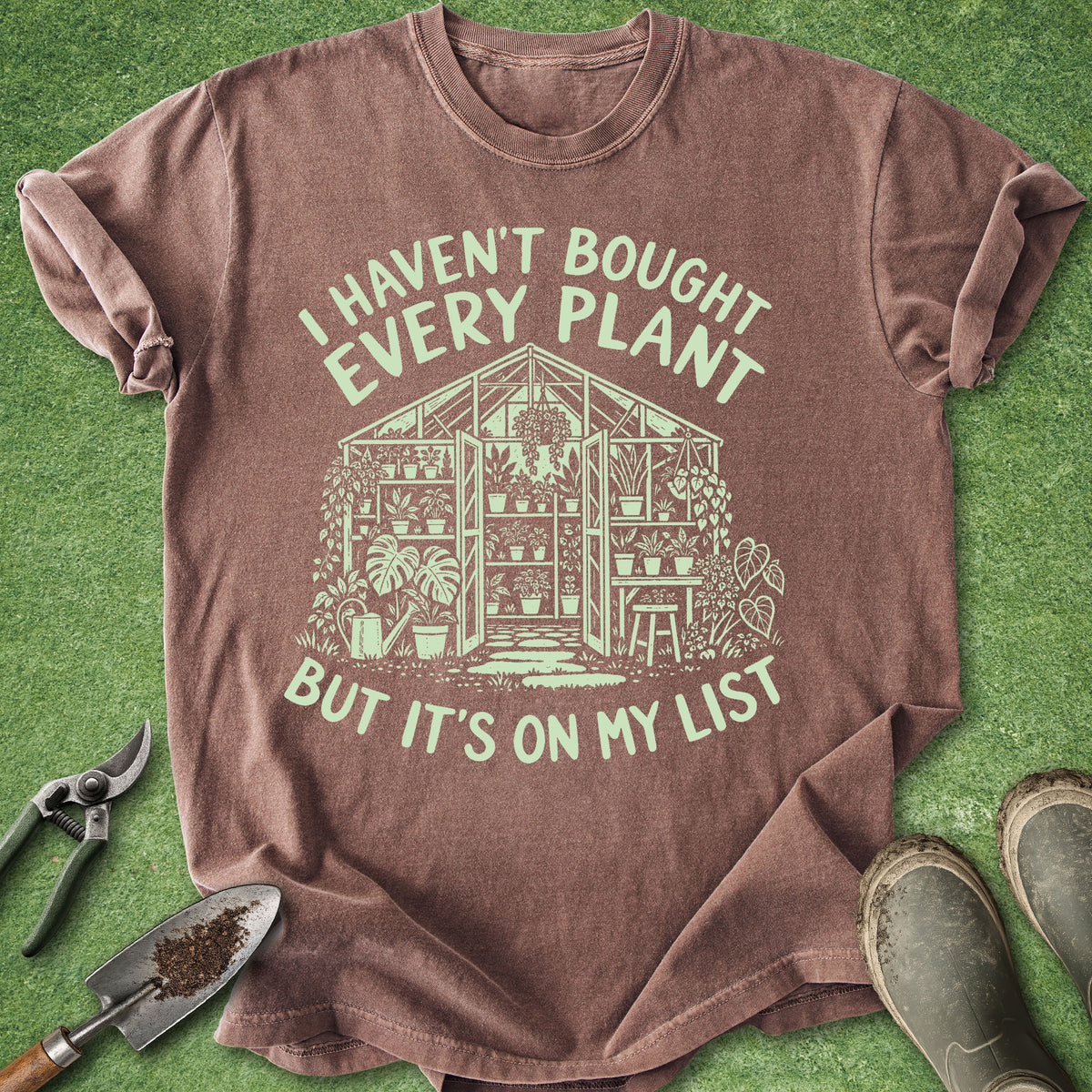 A brown short-sleeve graphic T-shirt laid flat on green grass-like background, with gardening tools and boots around it. The shirt has a large pale green printed design and text reading, 'I Haven't Bought Every Plant But It's On My List.