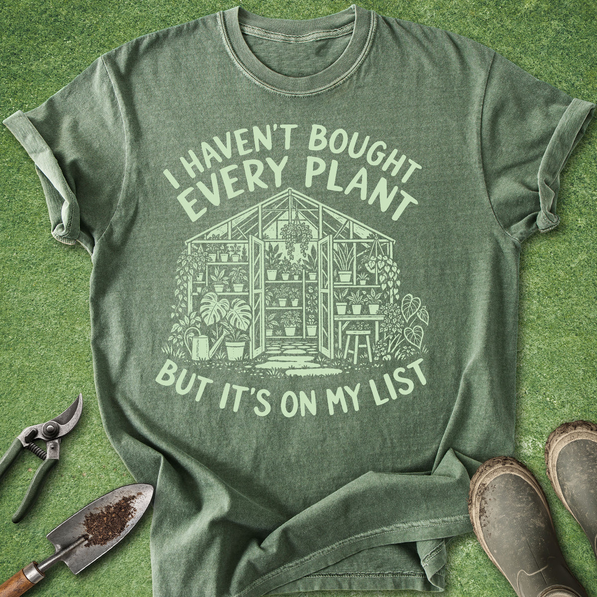 An olive green graphic t-shirt laid flat on a green grass-like surface, surrounded by gardening props including pruning shears, a trowel, and boots. The shirt features a light green graphic of a greenhouse with plants and the text 'I HAVEN'T BOUGHT EVERY PLANT BUT IT'S ON MY LIST'.