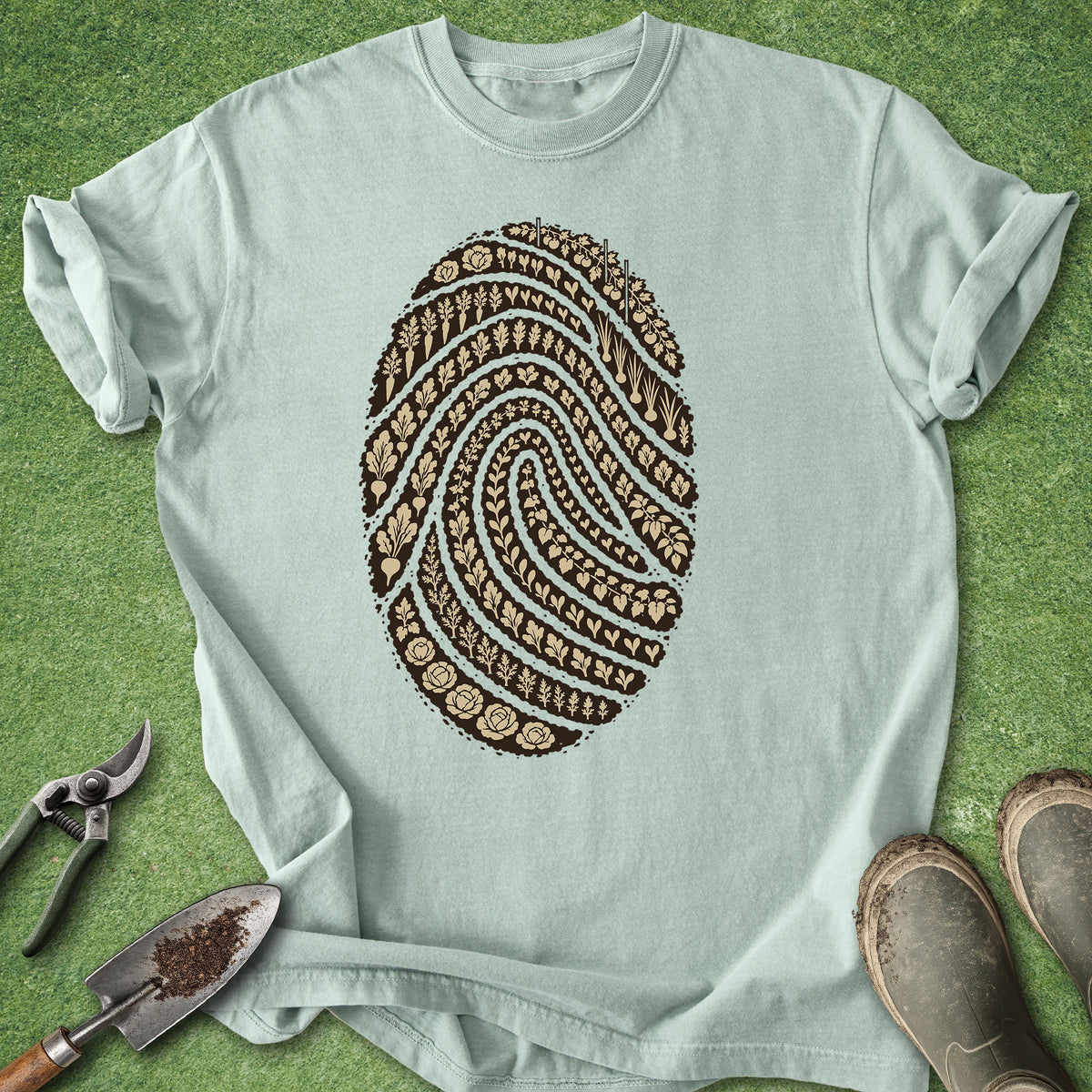 A light mint green t-shirt laid flat on a green grass-like surface, featuring a large brown fingerprint graphic filled with soil and gardening tool details. A pair of pruning shears, a small trowel, and gardening boots are placed around the shirt as styling props.