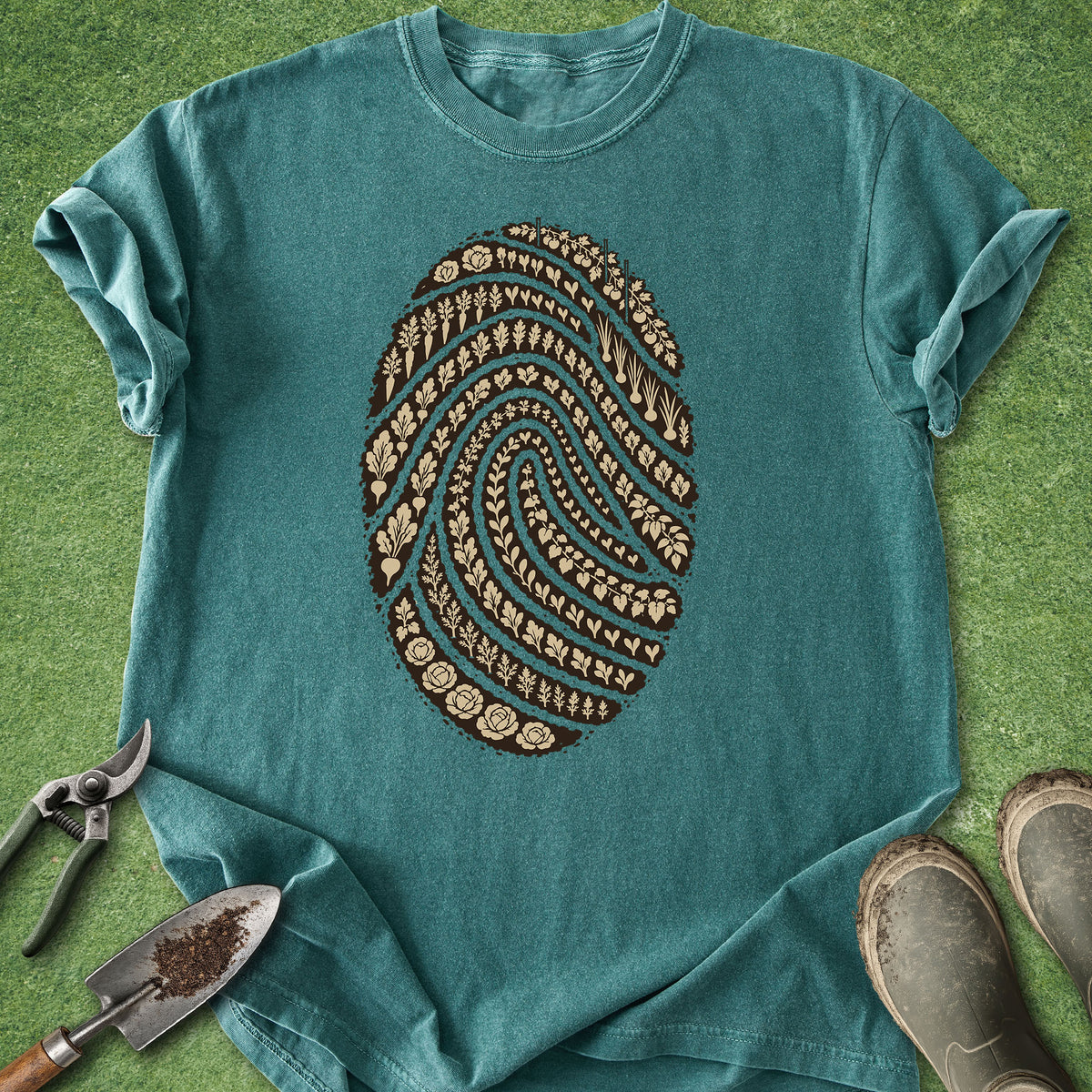 A teal short-sleeve t-shirt laid flat on a green grass-like surface, featuring a large brown fingerprint graphic filled with small floral and leaf motifs. Gardening tools and muddy boots are placed around the shirt as styling props.