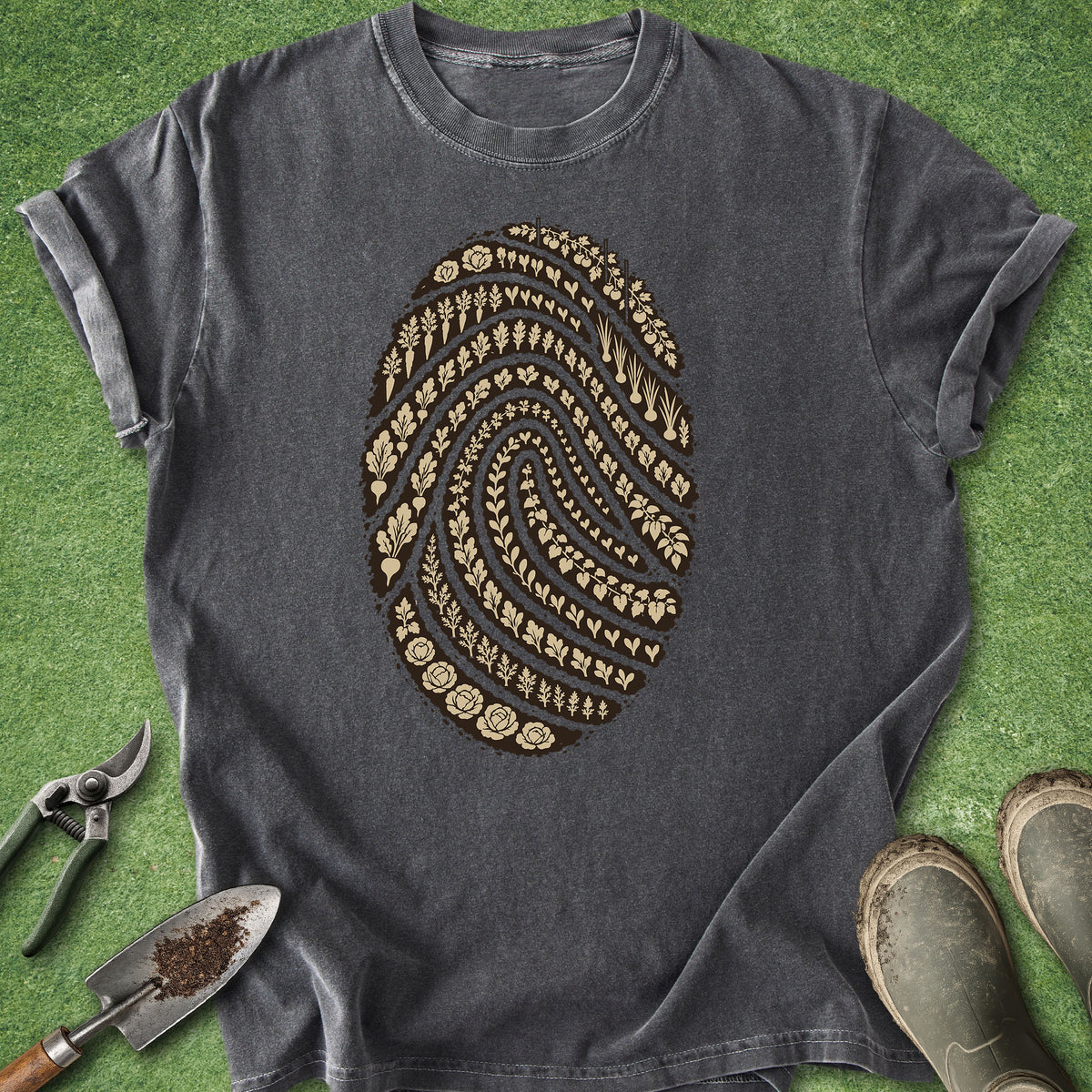 A dark gray crewneck t-shirt with a large brown fingerprint graphic made of gardening tools and seeds, laid flat on green grass with gardening props like a trowel, pruning shears, and boots.