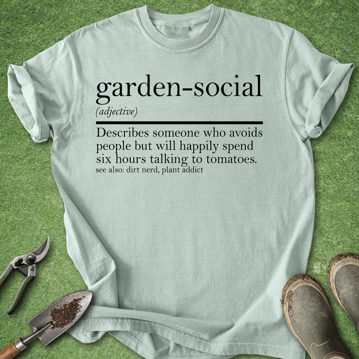 A light sage green graphic t-shirt laid flat on a green grass-like surface, styled with gardening tools (shears, trowel) and work boots. The t-shirt features a black printed dictionary-style definition of the term 'garden-social'.