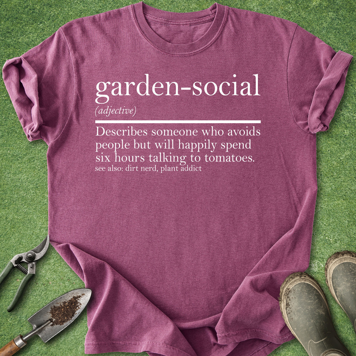 A heathered mauve-purple t-shirt with a white printed dictionary-style definition reading 'garden-social' and a humorous descriptor about avoiding people while spending time with tomatoes. The shirt is laid flat on green grass with gardening props including pruning shears, a trowel, and boots around it.