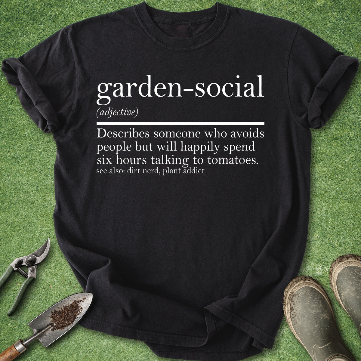 A black graphic t-shirt laid flat on green grass-like artificial turf, with gardening tools and work boots around it. The shirt has white printed text defining the phrase 'garden-social'.