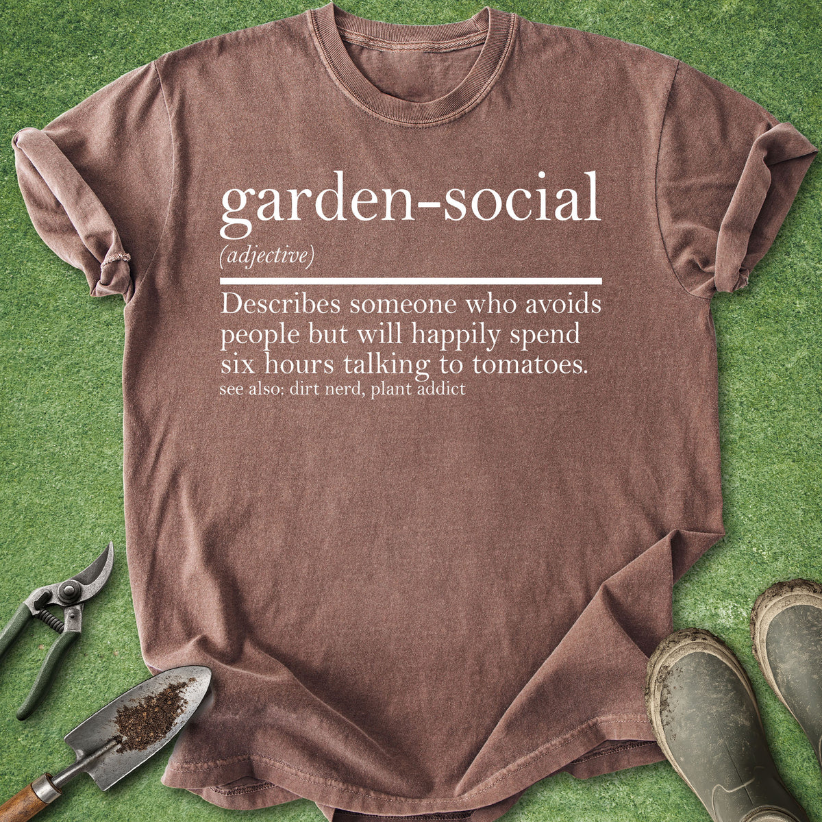 A brown graphic t-shirt laid flat on green grass with gardening tools and boots around it. The shirt has a white printed dictionary-style definition reading 'garden-social' with a humorous explanatory phrase below.