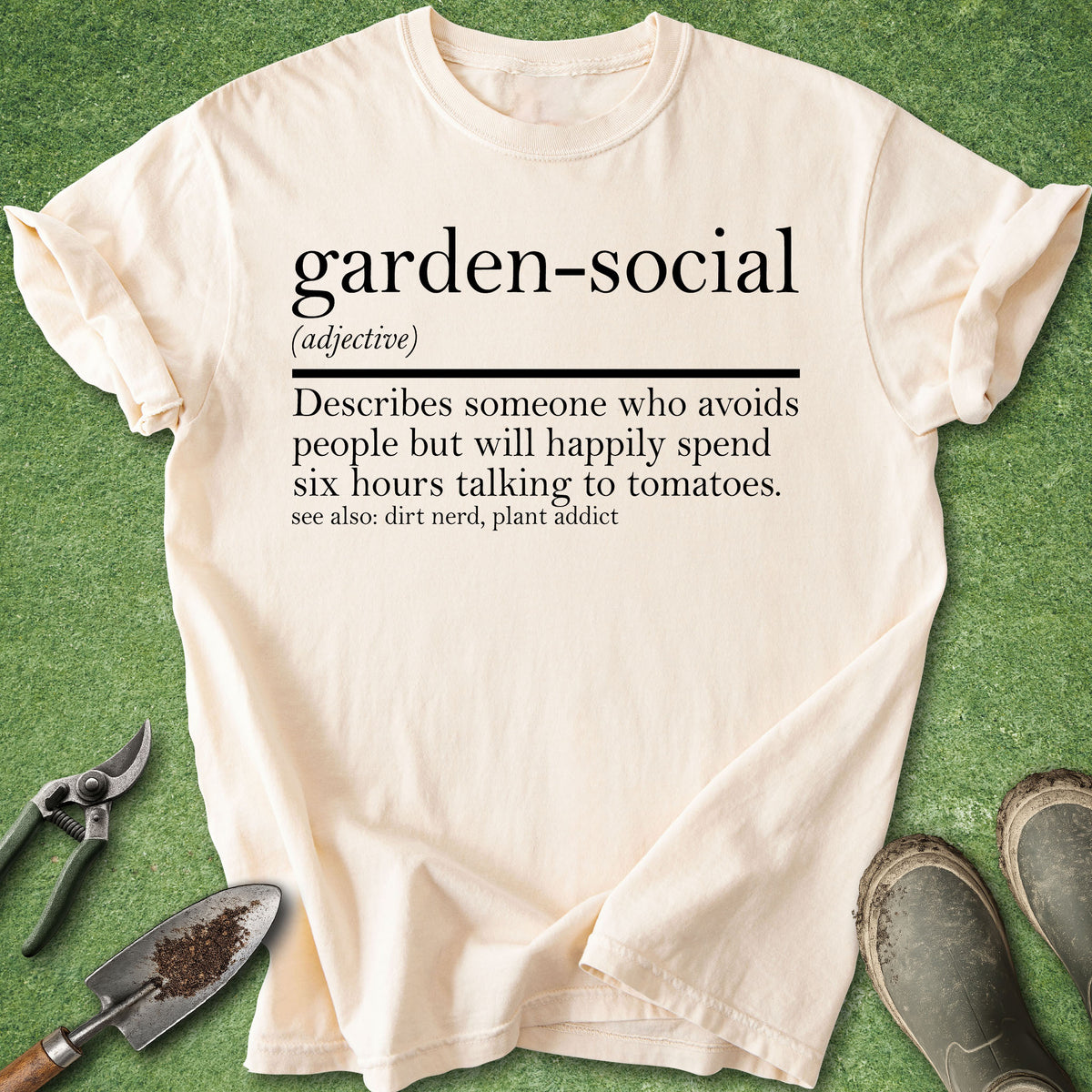 A cream-colored graphic t-shirt laid flat on green grass, surrounded by gardening props including pruning shears, a trowel, and gardening boots. The t-shirt features a humorous dictionary-style definition of the term 'garden-social'.