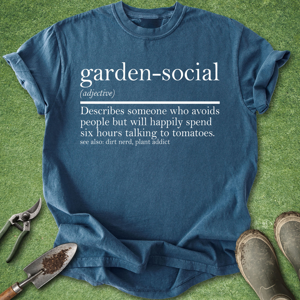 A blue graphic t-shirt laid flat on green grass-like background, surrounded by gardening tools and boots. The shirt has white printed text defining the term 'garden-social'.