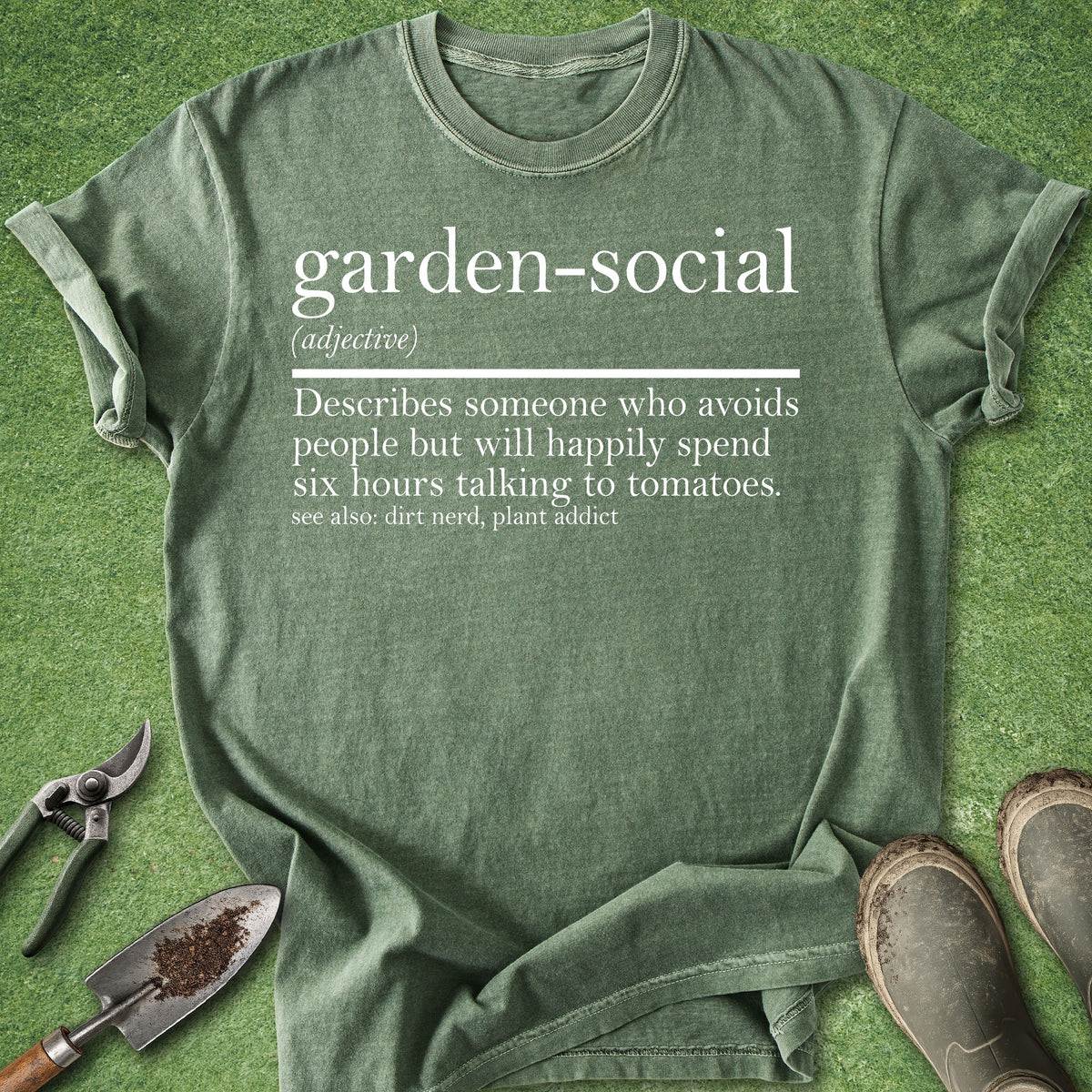 An olive green graphic t-shirt laid flat on a green grass-like surface, styled with gardening tools and muddy boots. The t-shirt features a white printed dictionary-style definition of the term 'garden-social'.