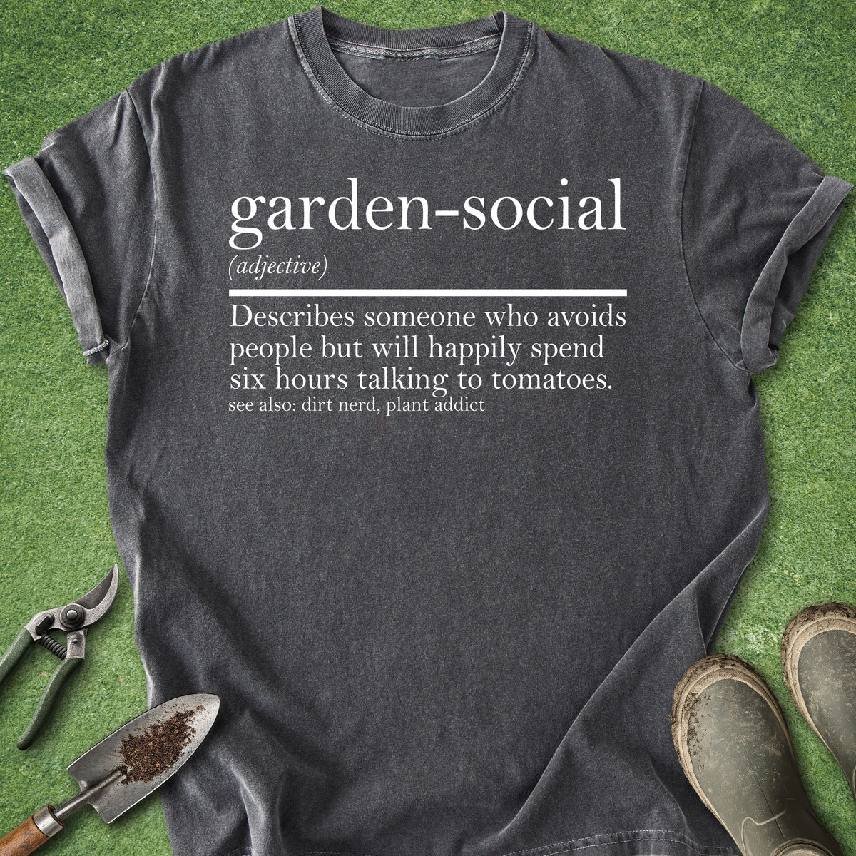A dark charcoal gray graphic t-shirt laid flat on green grass with gardening props, including pruning shears, a small hand trowel, and muddy boots. The shirt features a white printed dictionary-style definition of the term 'garden-social'.