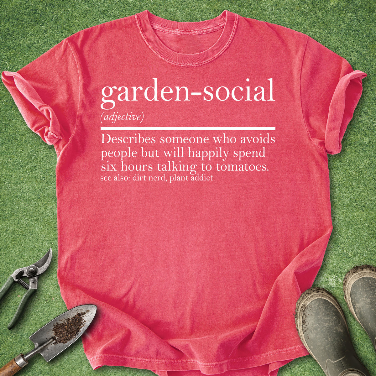 A coral pink graphic t-shirt laid flat on green grass, surrounded by gardening props including pruning shears, a small trowel, and work boots. The shirt features a white printed dictionary-style definition of the word 'garden-social'.