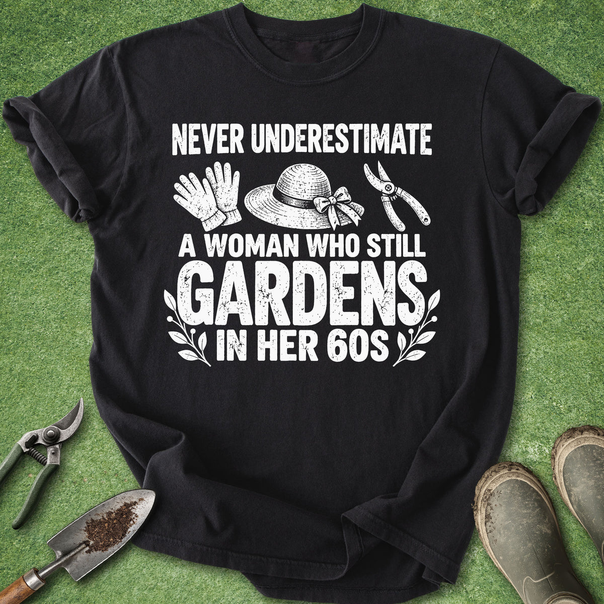 A black short-sleeve graphic T-shirt laid flat on green grass with gardening props around it. The shirt has a large white printed slogan reading 'NEVER UNDERESTIMATE A WOMAN WHO STILL GARDENS IN HER 60S' with small illustrations of gardening tools and a hat.