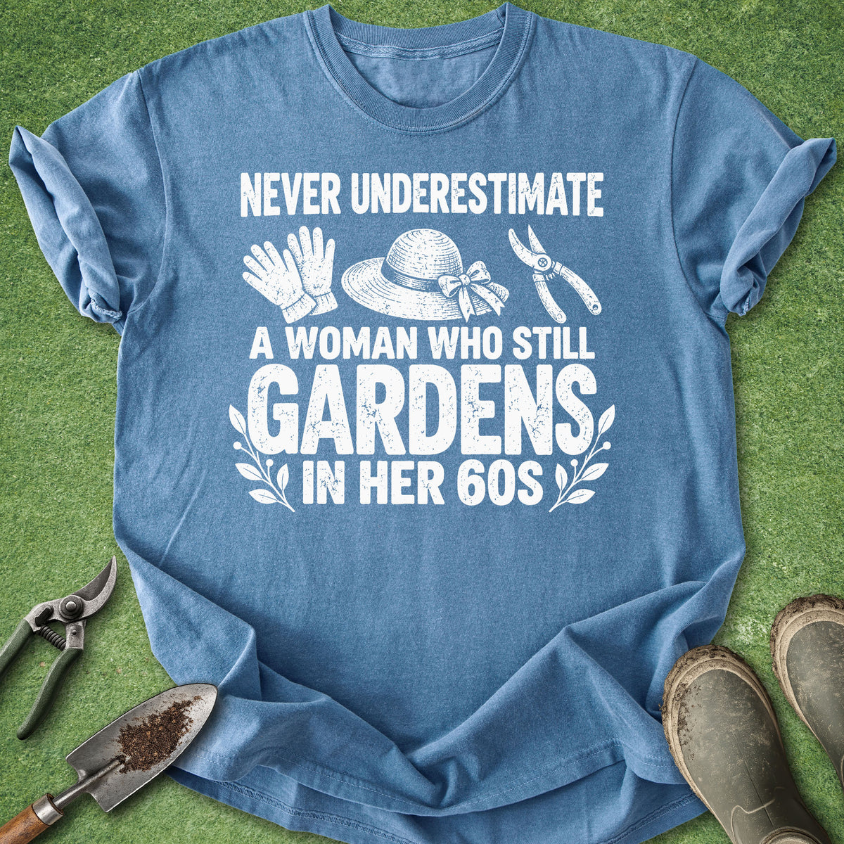 A heathered blue short-sleeve t-shirt with a white graphic print that reads 'NEVER UNDERESTIMATE A WOMAN WHO STILL GARDENS IN HER 60S' with illustrations of gardening gloves, a hat, shears, and a trowel. The shirt is laid flat on green grass-like material with gardening tools and boots around it.