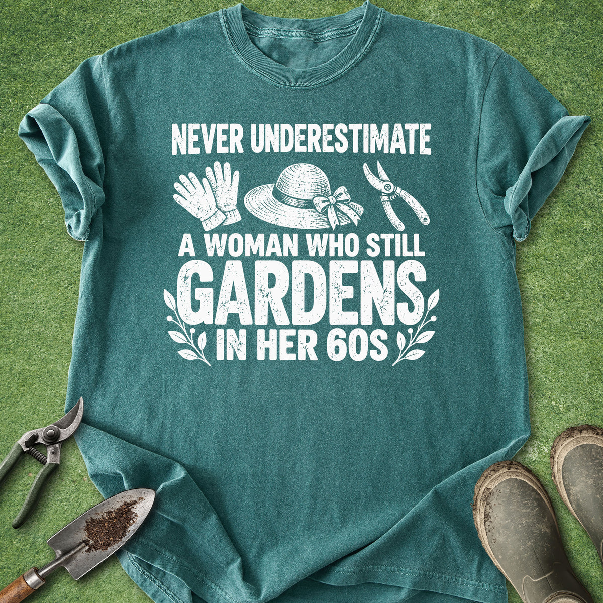 A teal graphic t-shirt laid flat on a green grass-like surface, surrounded by gardening props including pruning shears, a trowel, gloves, a hat, and boots. The shirt features a white distressed text design that reads 'NEVER UNDERESTIMATE A WOMAN WHO STILL GARDENS IN HER 60S' with small gardening icon illustrations.