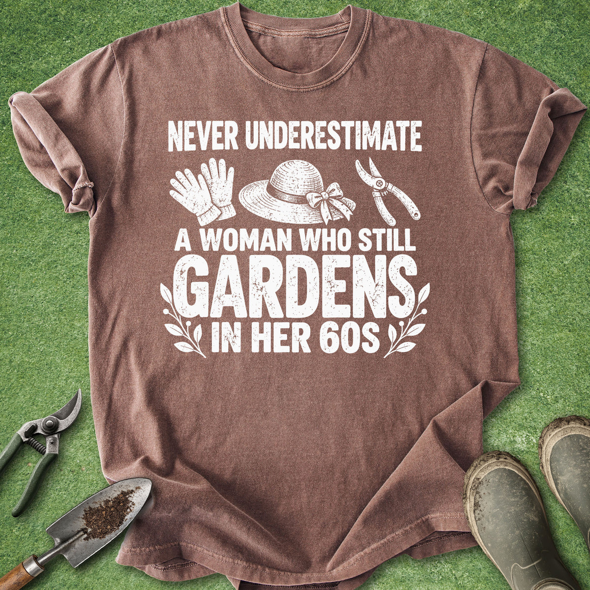 A brown short-sleeve graphic T-shirt laid flat on green artificial grass, surrounded by gardening props including pruning shears, a trowel, gloves, a hat, and boots. The shirt has a large white text print reading “NEVER UNDERESTIMATE A WOMAN WHO STILL GARDENS IN HER 60S” with small gardening icon illustrations.