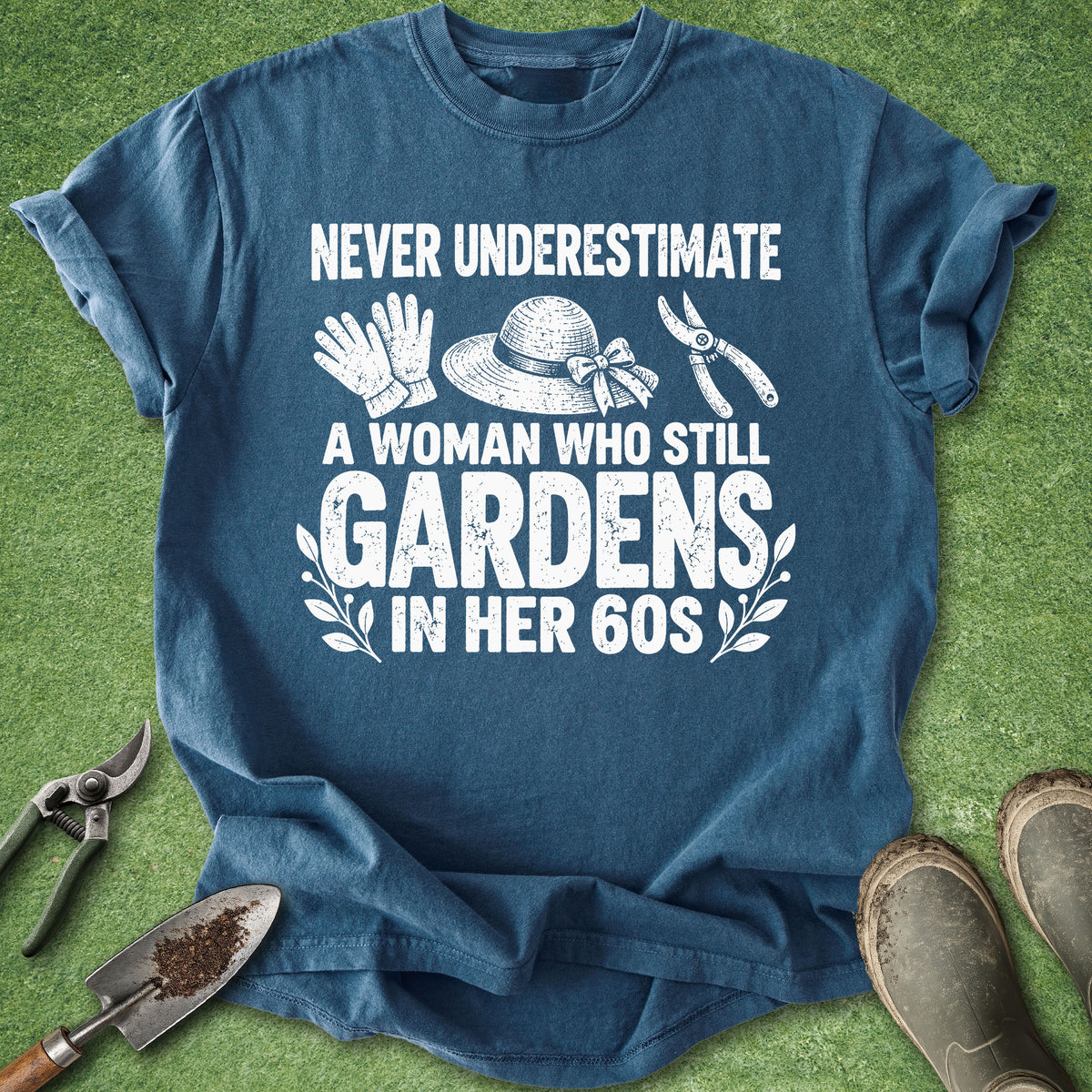 A blue short-sleeve graphic T-shirt laid flat on green grass with gardening props around it. The shirt has white text reading 'NEVER UNDERESTIMATE A WOMAN WHO STILL GARDENS IN HER 60S' with illustrated gloves, a hat, pruning shears, and leaf motifs.
