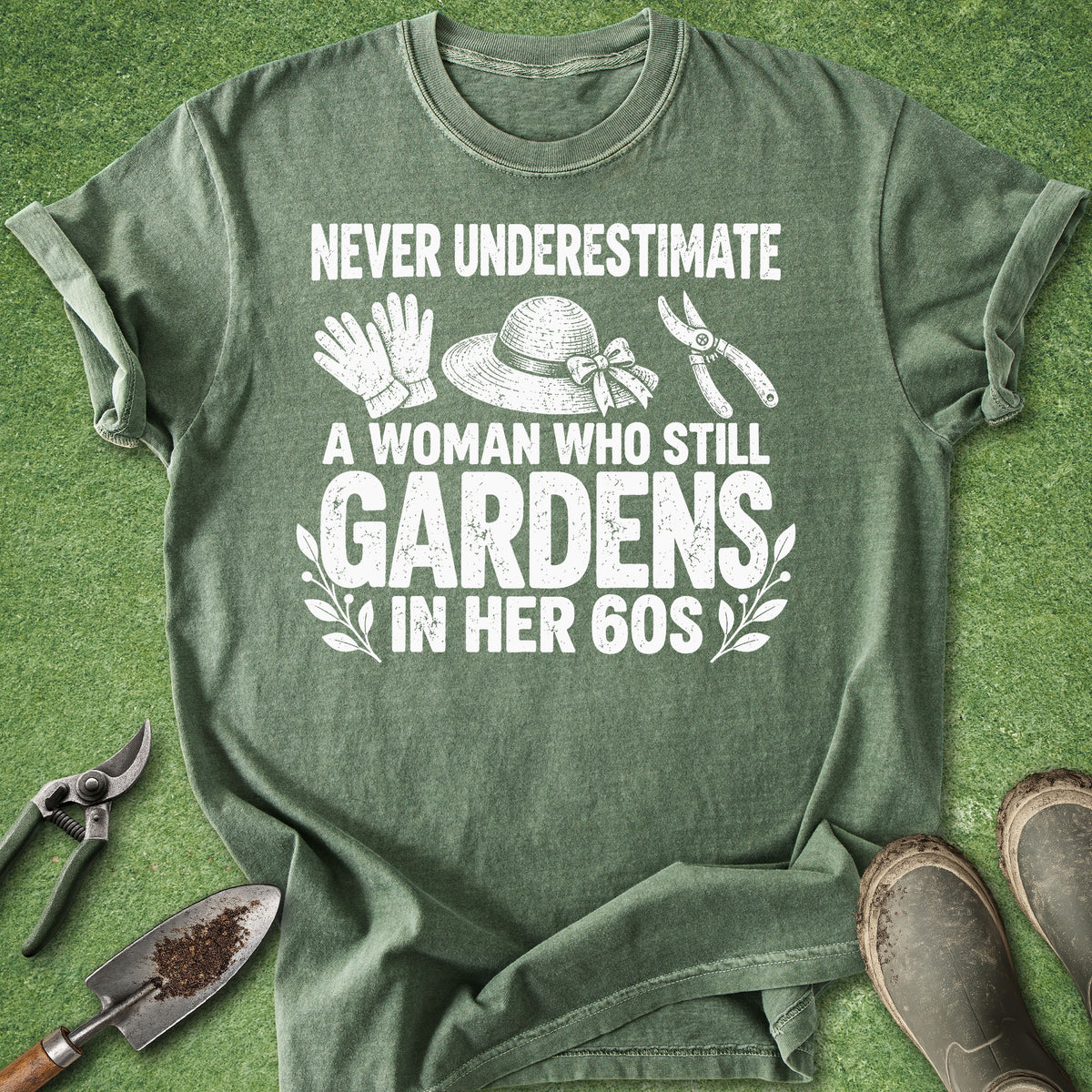 An olive green graphic t-shirt laid flat on a green grass-like surface, surrounded by gardening props including pruning shears, a trowel, a small shovel, and work boots. The shirt features a white graphic print with gardening tools and the text 'NEVER UNDERESTIMATE A WOMAN WHO STILL GARDENS IN HER 60S'.