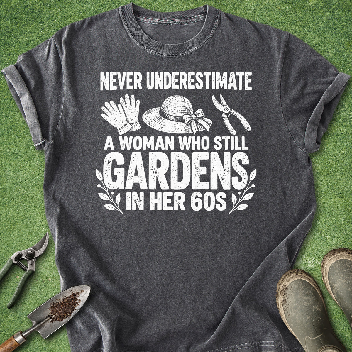A dark gray graphic t-shirt laid flat on green artificial grass, surrounded by gardening props including pruning shears, a trowel, gloves, a hat, and boots. The shirt features a white distressed text graphic that reads 'NEVER UNDERESTIMATE A WOMAN WHO STILL GARDENS IN HER 60S' with illustrations of gardening tools and a hat.