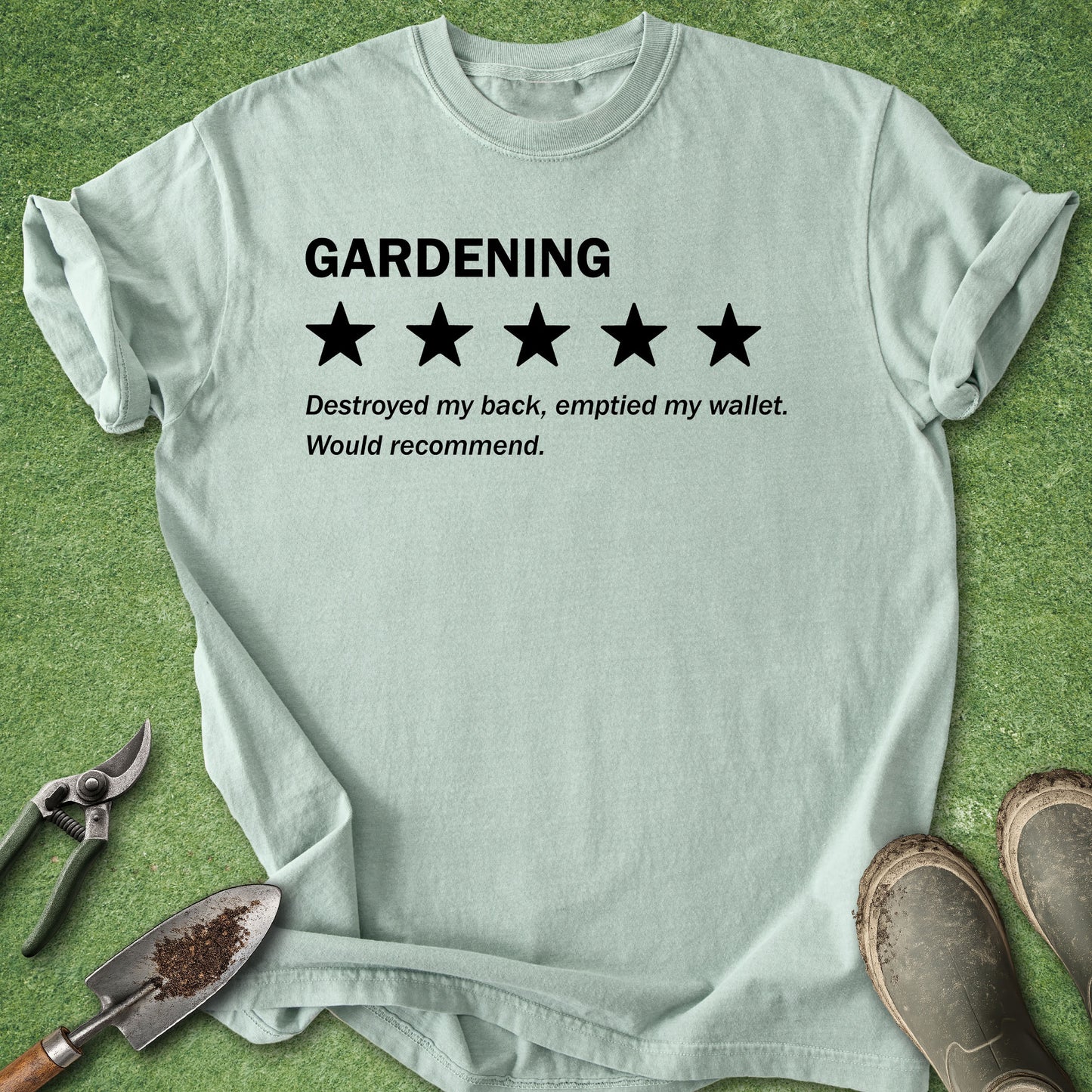 A light sage green graphic t-shirt laid flat on a green grass-like surface, with gardening tools and boots around it. The shirt has black text reading 'GARDENING' above a five-star rating graphic and a humorous review below.
