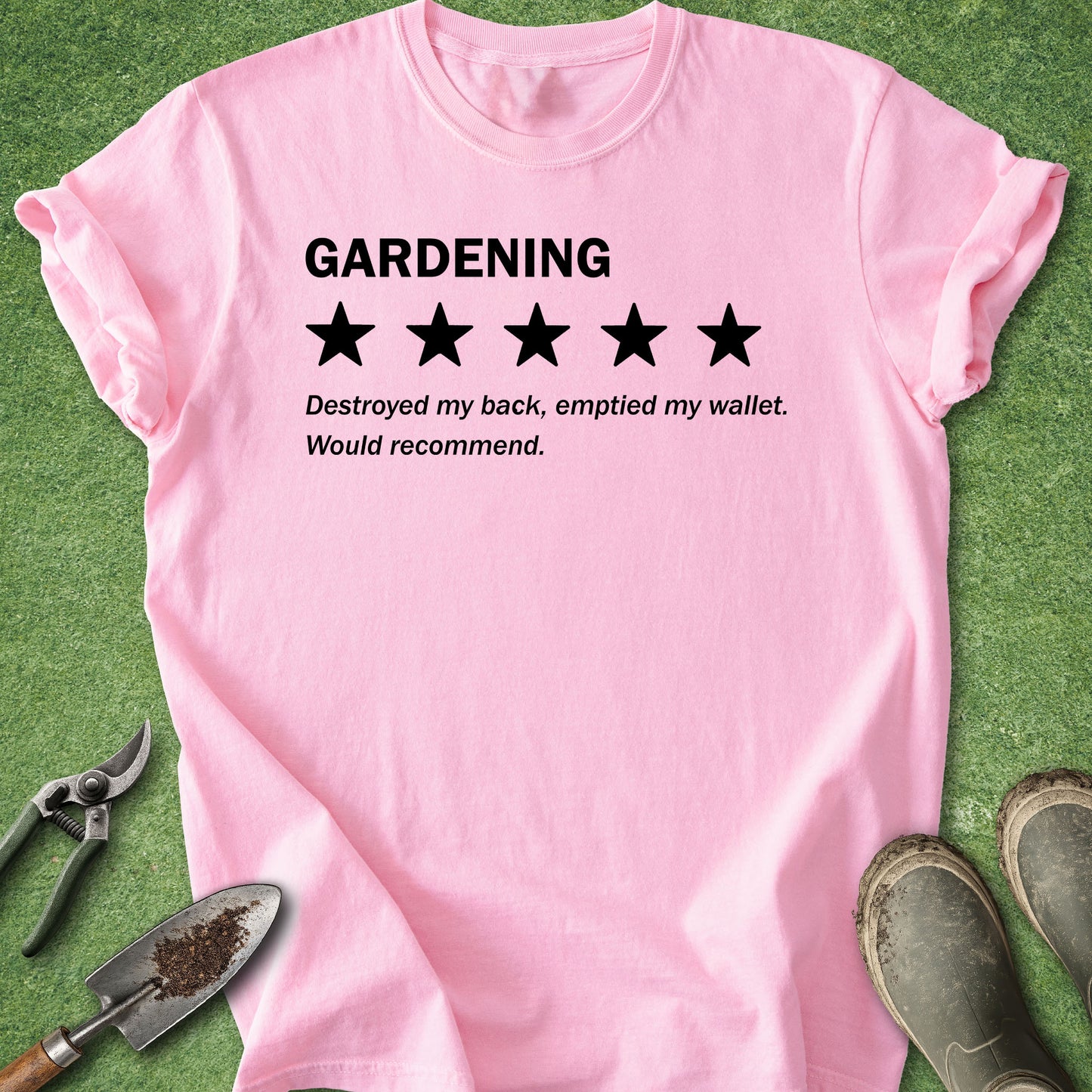 A pink short-sleeve t-shirt with a humorous gardening review graphic, laid flat on green grass with gardening tools and boots around it.