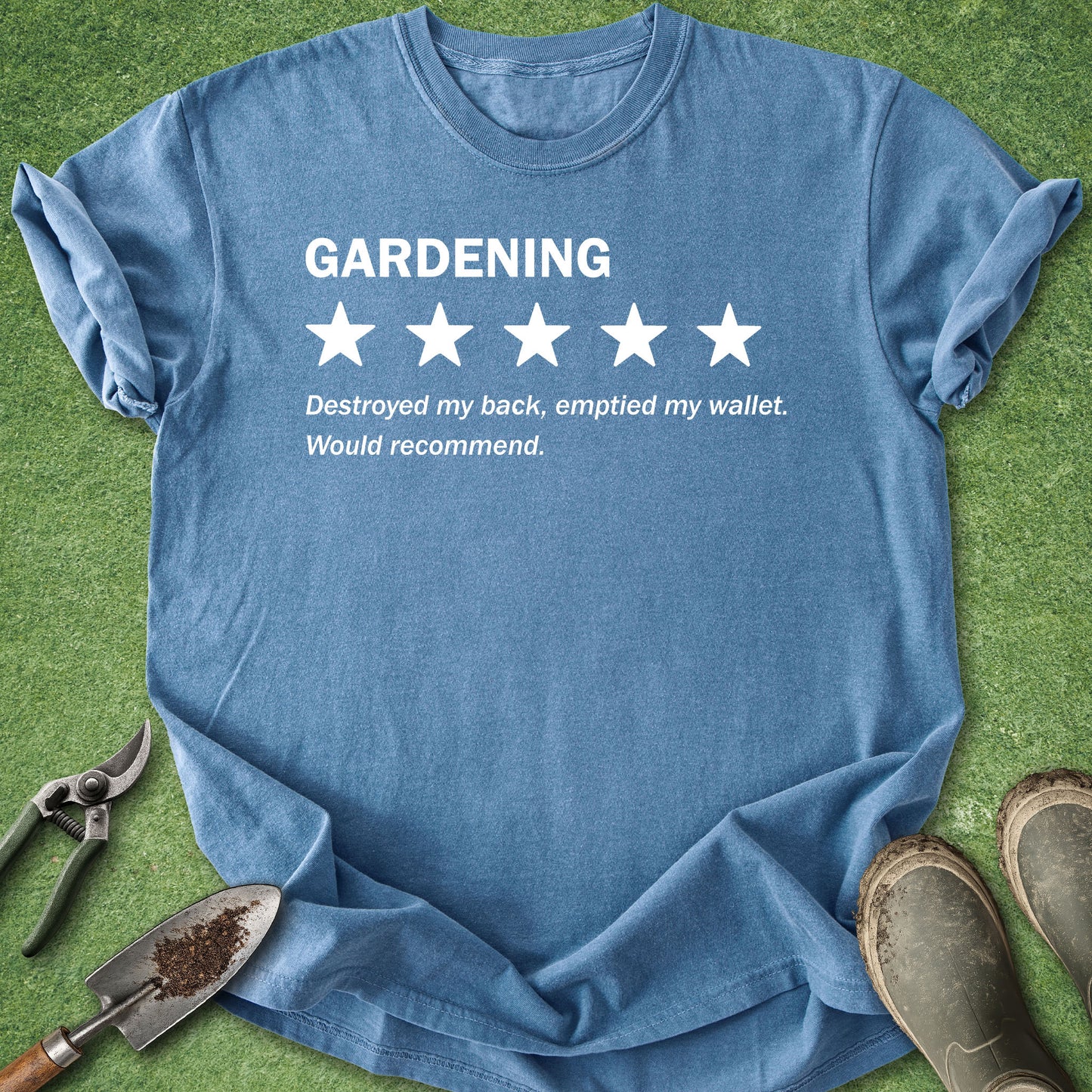 A blue short-sleeve graphic T-shirt laid flat on green grass with gardening props around it. The shirt has white text reading 'GARDENING' above five white stars and the phrase 'Destroyed my back, emptied my wallet.
