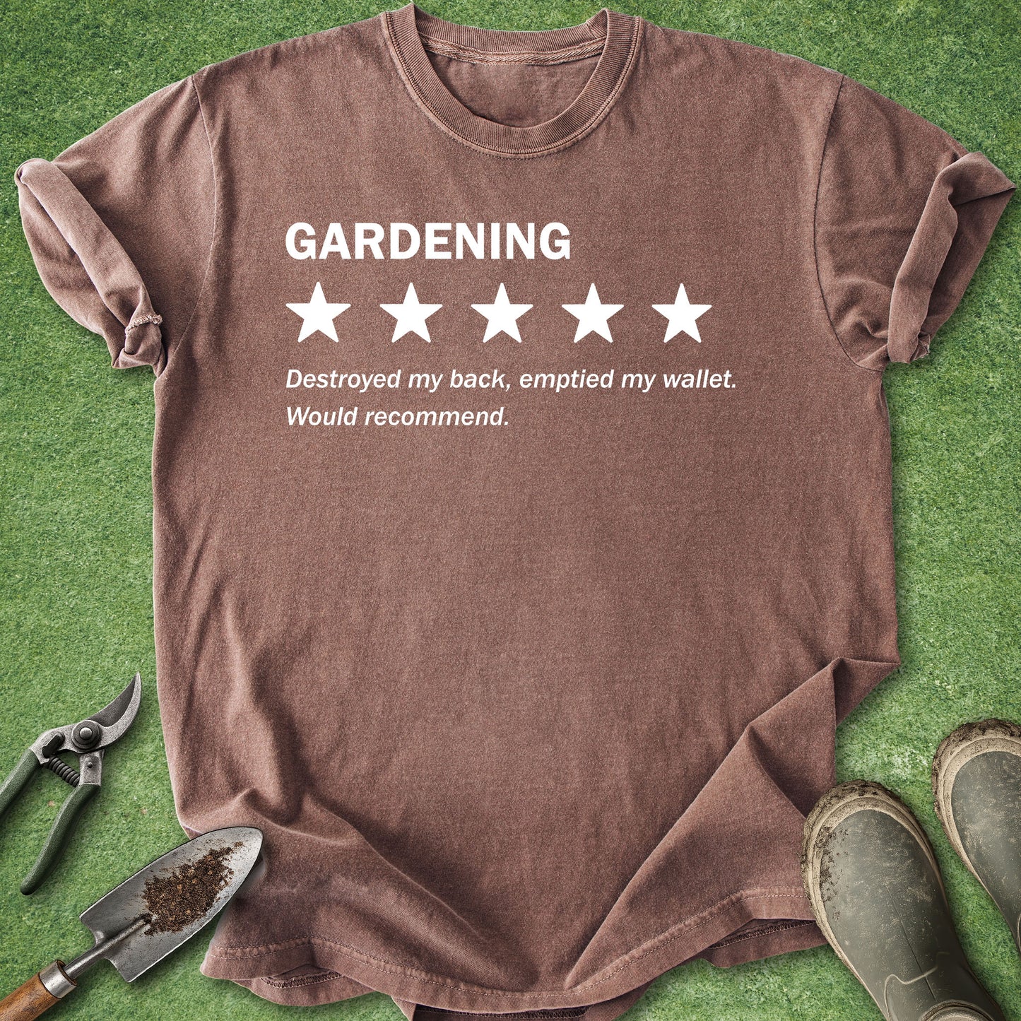 A brown short-sleeve t-shirt with a white graphic print reading 'GARDENING' above five stars and a humorous quote, laid flat on green grass with gardening tools and boots nearby.