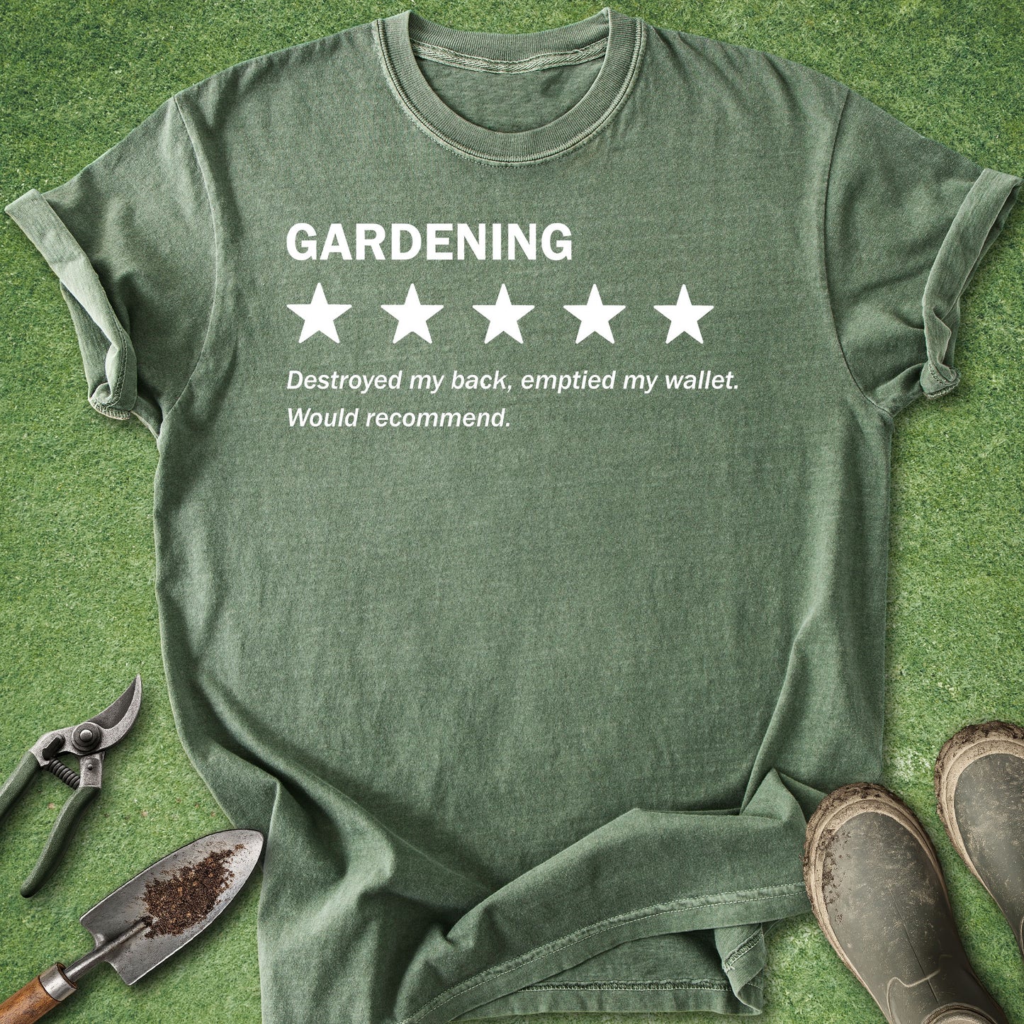 An olive green graphic t-shirt laid flat on a green grass background, styled with gardening tools (trowel, pruning shears) and muddy boots. The shirt features a white graphic that reads 'GARDENING' above a 1-star rating, with the text 'Destroyed my back, emptied my wallet.