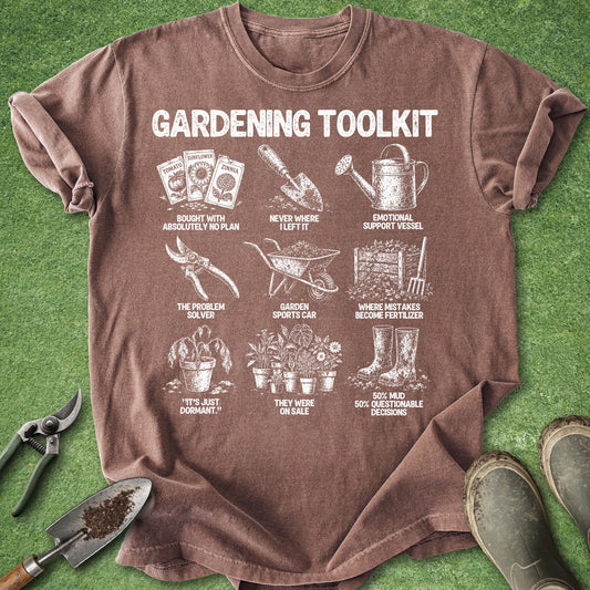 A brown short-sleeve graphic T-shirt laid flat on green grass-like background, with gardening-themed white illustrations and text including tools, pots, gloves, and boots. A pair of pruning shears, a small hand tool, and boots are placed around the shirt as styling props.