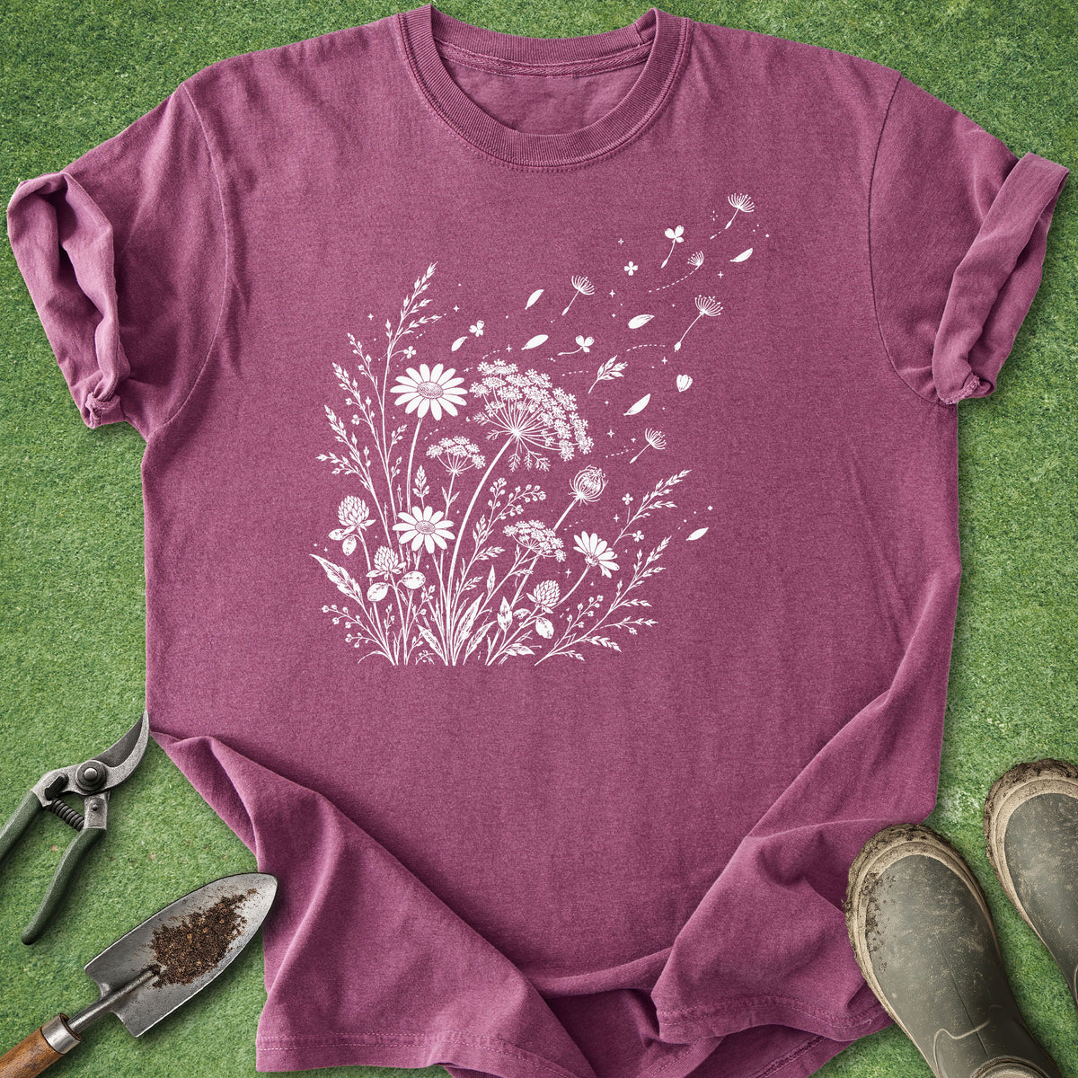 A heathered mauve-purple short-sleeve t-shirt with a white line-art graphic of wildflowers, laid flat on green grass with gardening tools and boots around it.