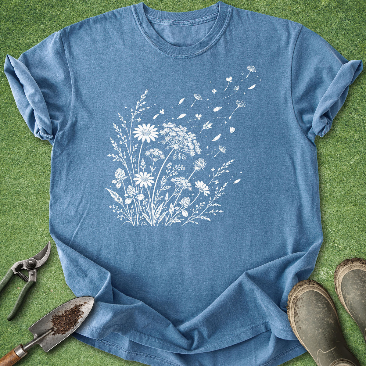 A blue short-sleeve crewneck t-shirt with a white dandelion and wildflower graphic, laid flat on green grass with gardening props including pruning shears, a trowel, and boots.