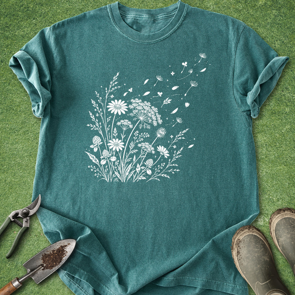 A teal short-sleeve crewneck t-shirt with a white danTeal green shirt featuring a delicate white wildflower botanical illustration with daisies, clover, grasses, petals and seeds blowing in the breeze.delion and wildflower graphic on the front, styled flat on green grass with gardening tools and boots.